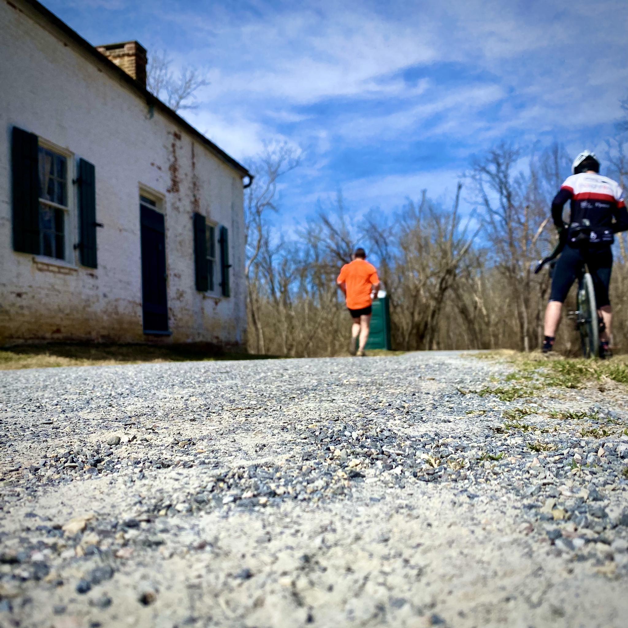 Two people on gravel path near old white building, bright day.