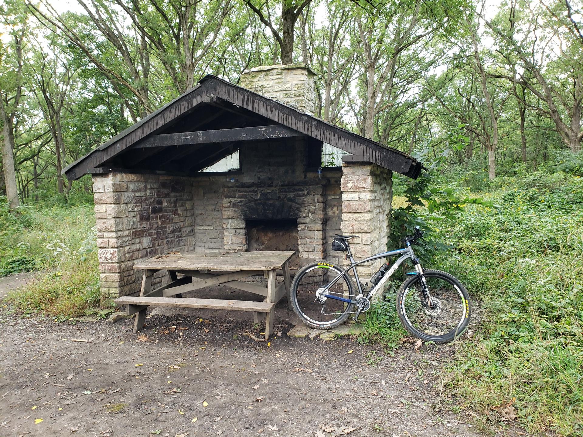 Stone shelter with picnic table and bicycle in a wooded area.