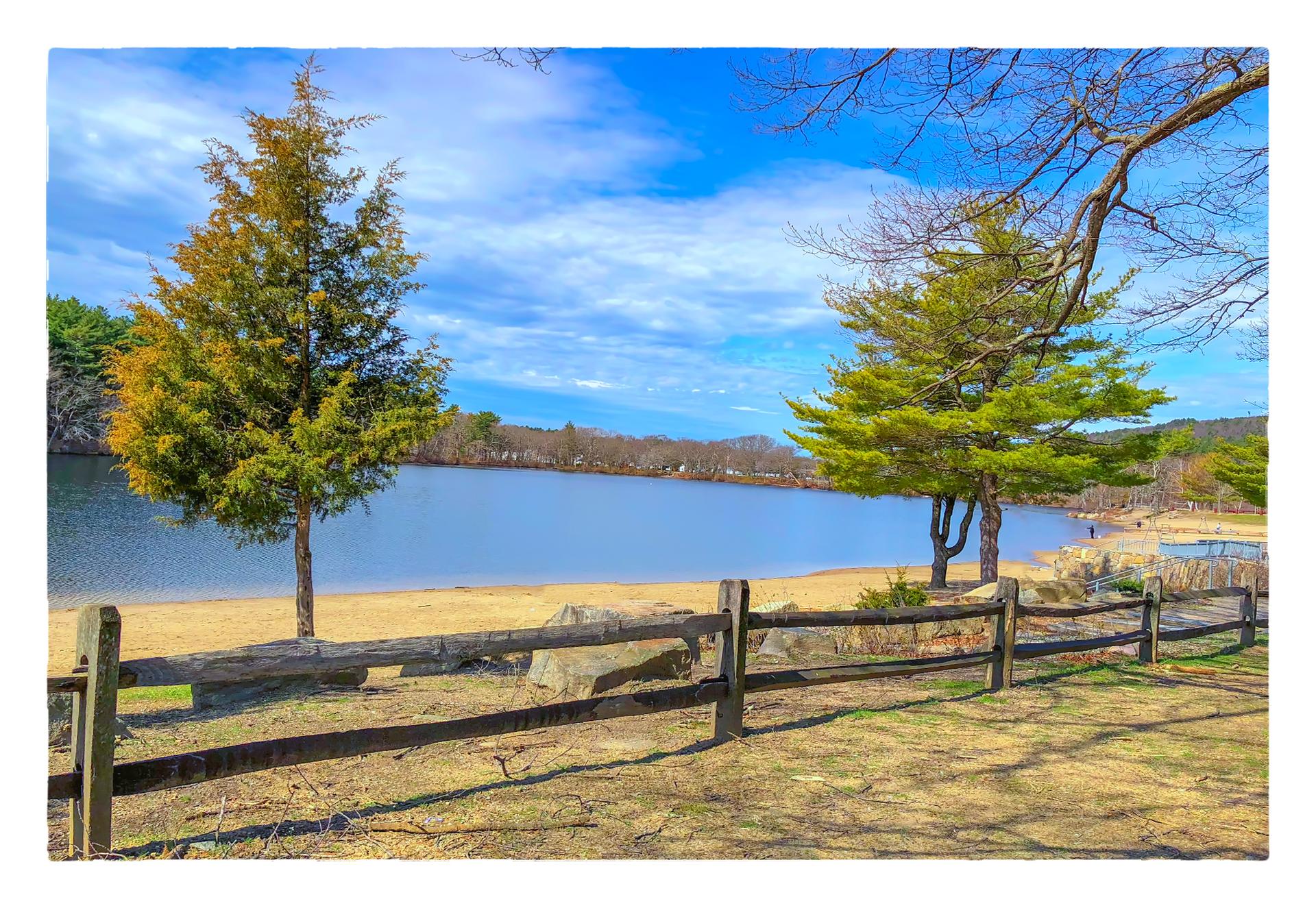 A sunny lake view with a sandy beach and wooden fence.