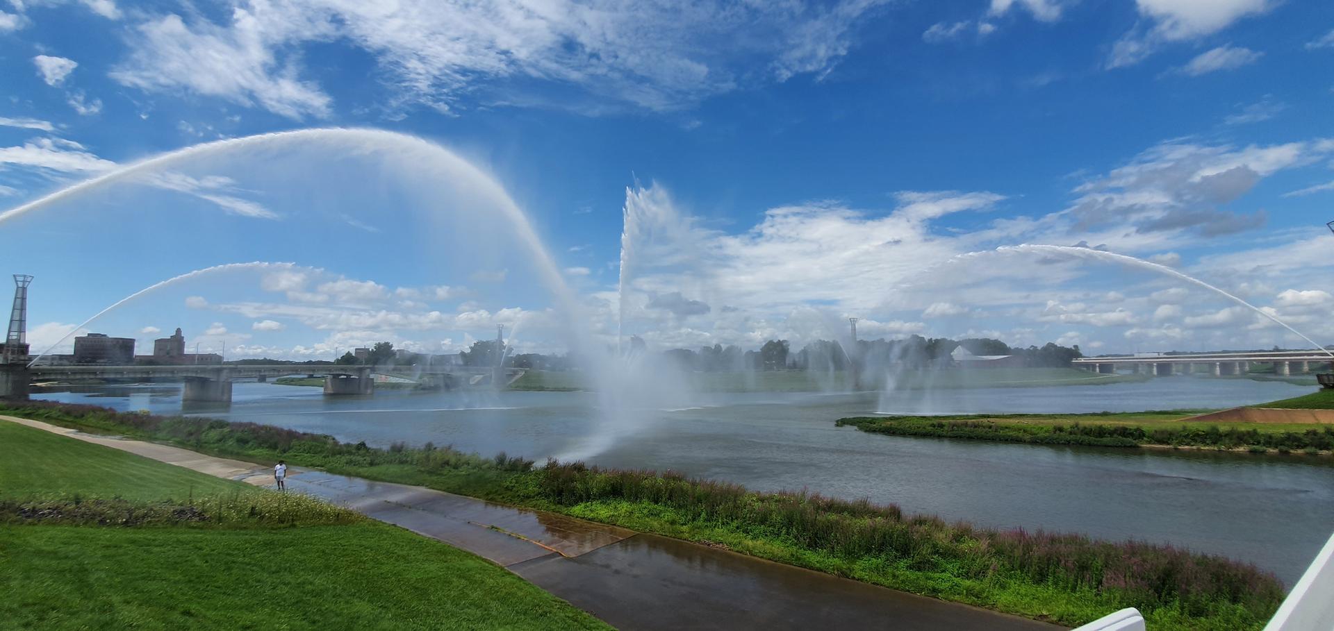 Large water fountains spray over a wide river under a bright blue sky.