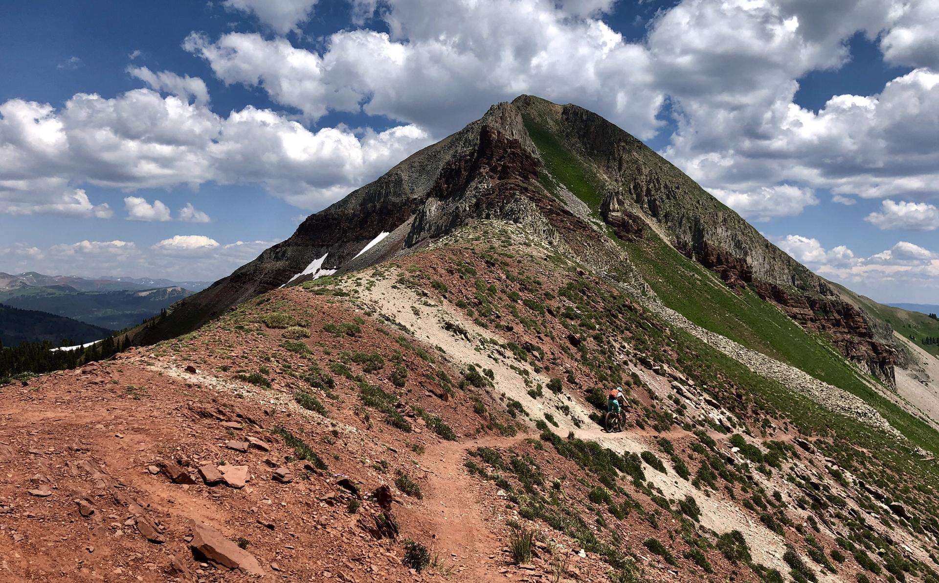 Mountain biker on a narrow trail ascending a rocky, green peak under a cloudy sky.