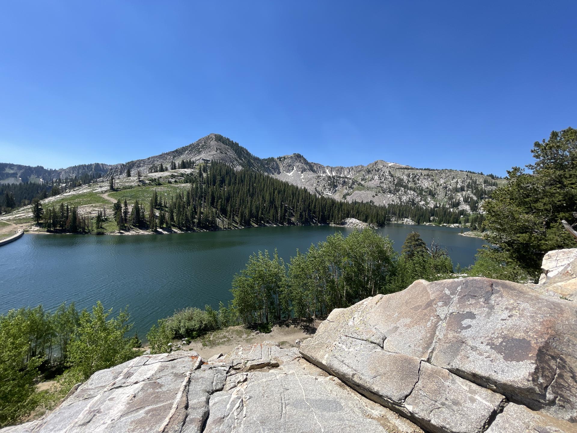 A clear blue sky over a mountain lake, surrounded by green forests and rocky peaks.