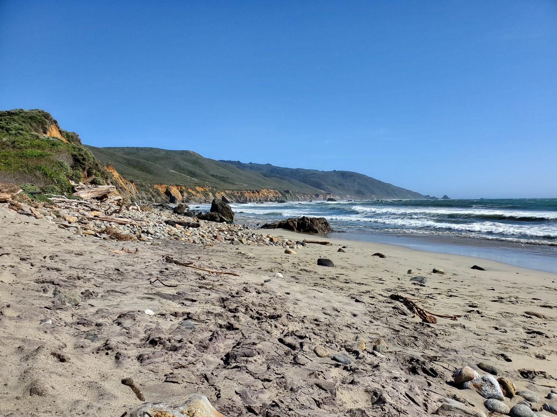Sunny rocky beach with ocean waves and a steep, green hillside.