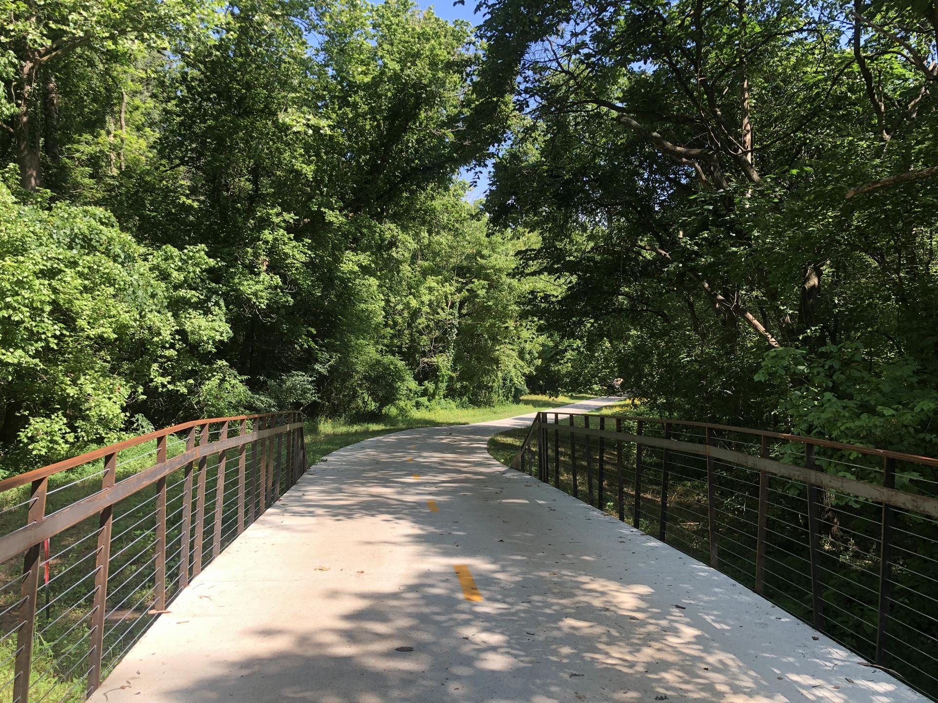 A paved trail bridge extends into a lush green forest on a sunny day.