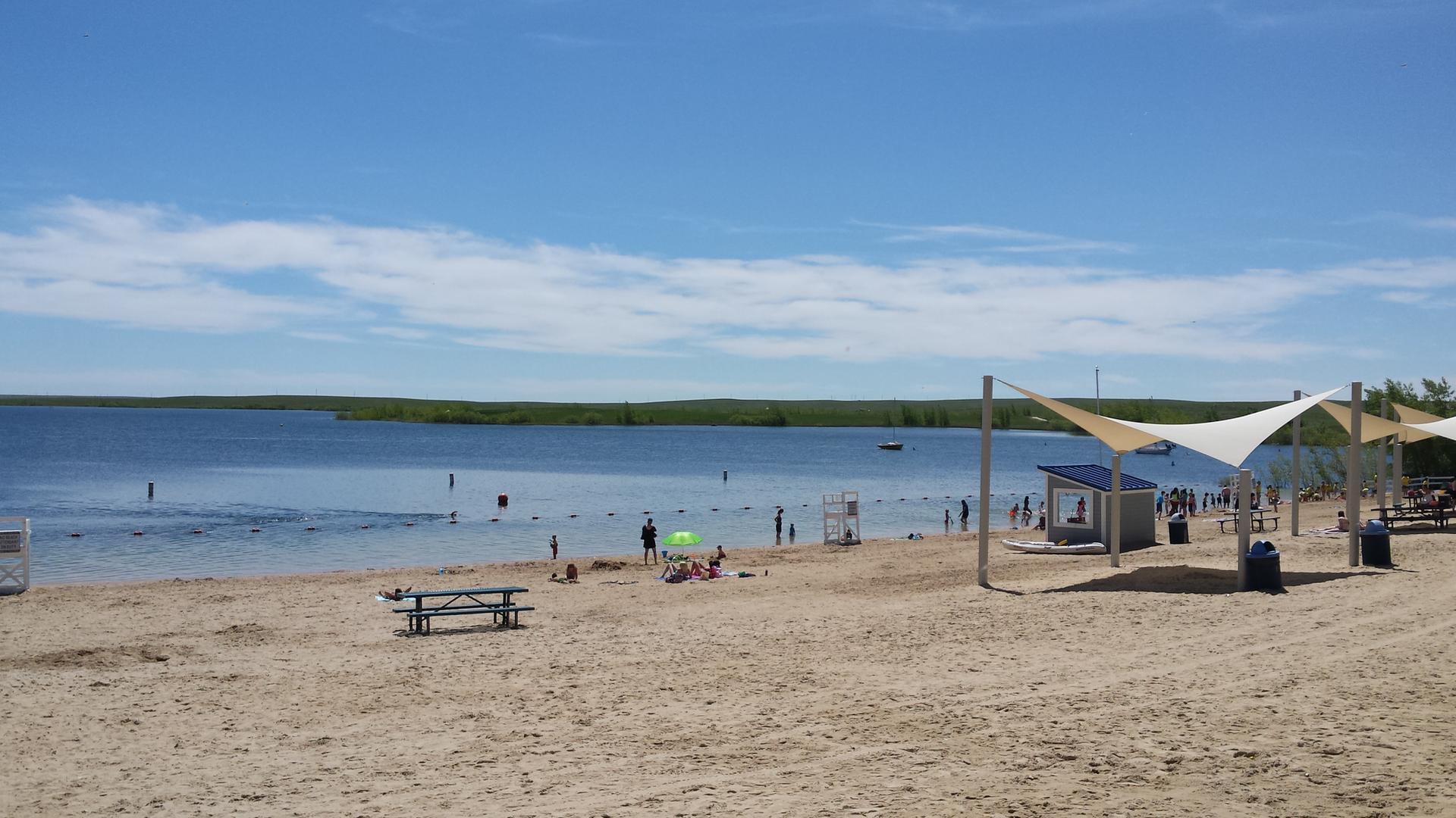 Sandy beach with people swimming and relaxing under shade sails on a sunny day.