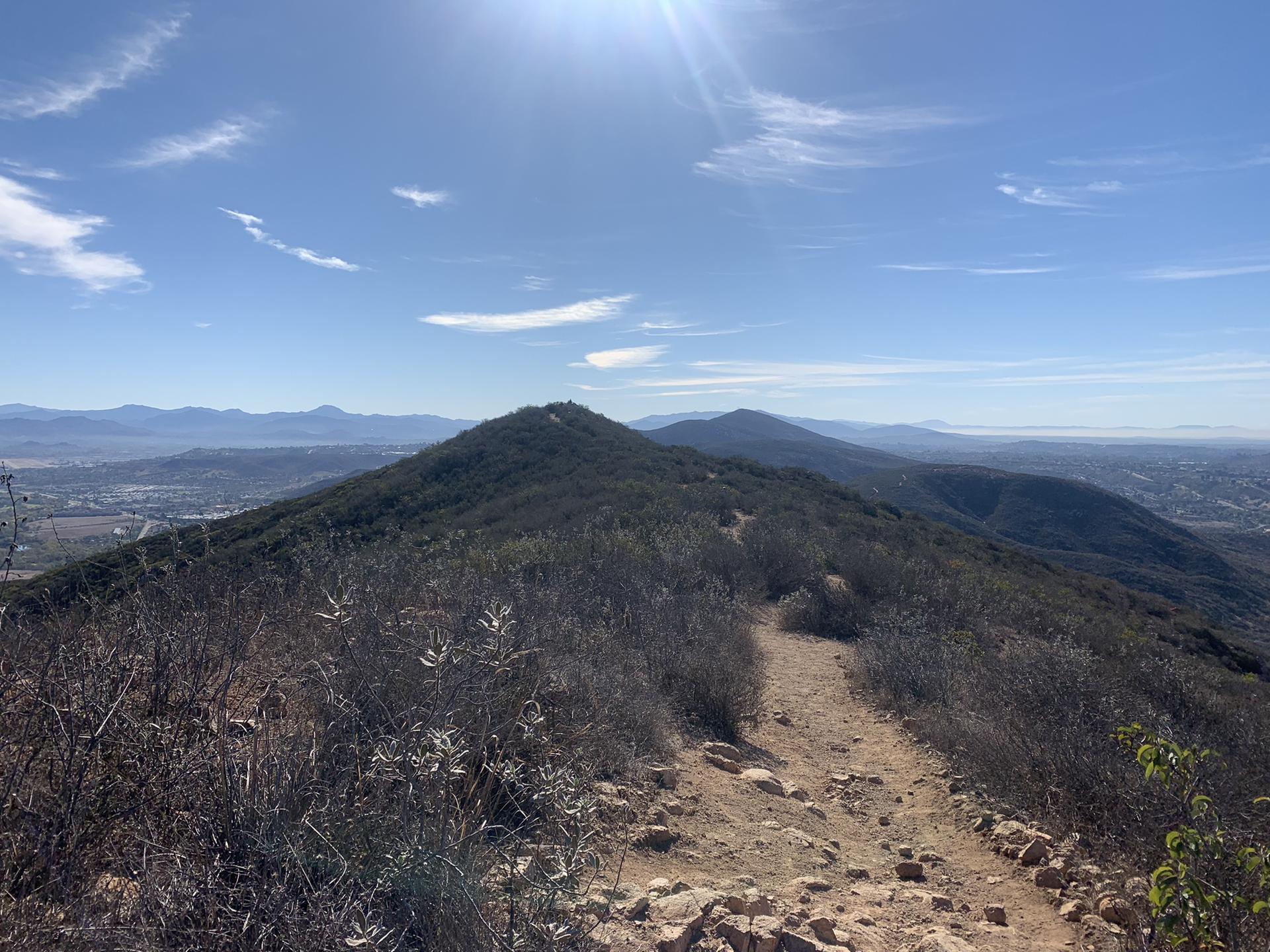 Narrow dirt trail on a mountain ridge under a bright, sunny blue sky.