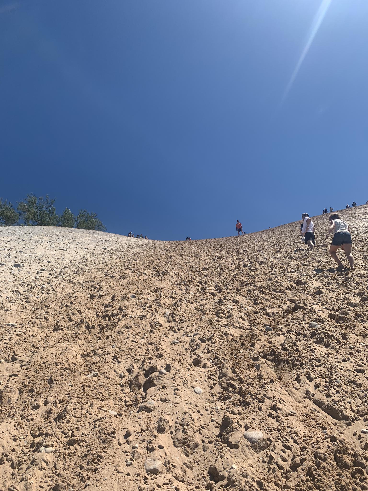People climb a very steep, sunny sand dune under a clear sky.