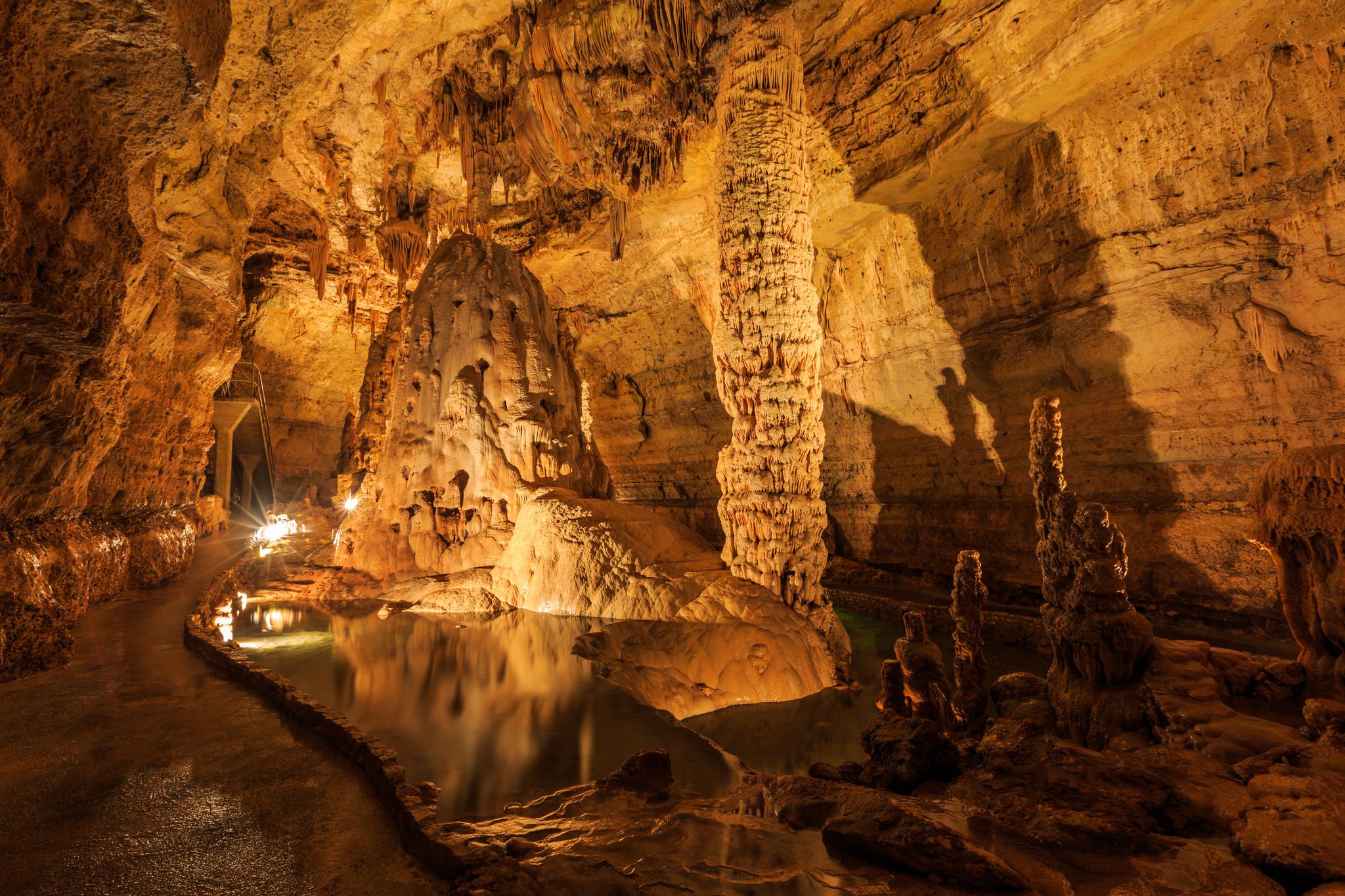 Warmly lit cave interior with large stalagmites, stalactites, and a reflective pool.