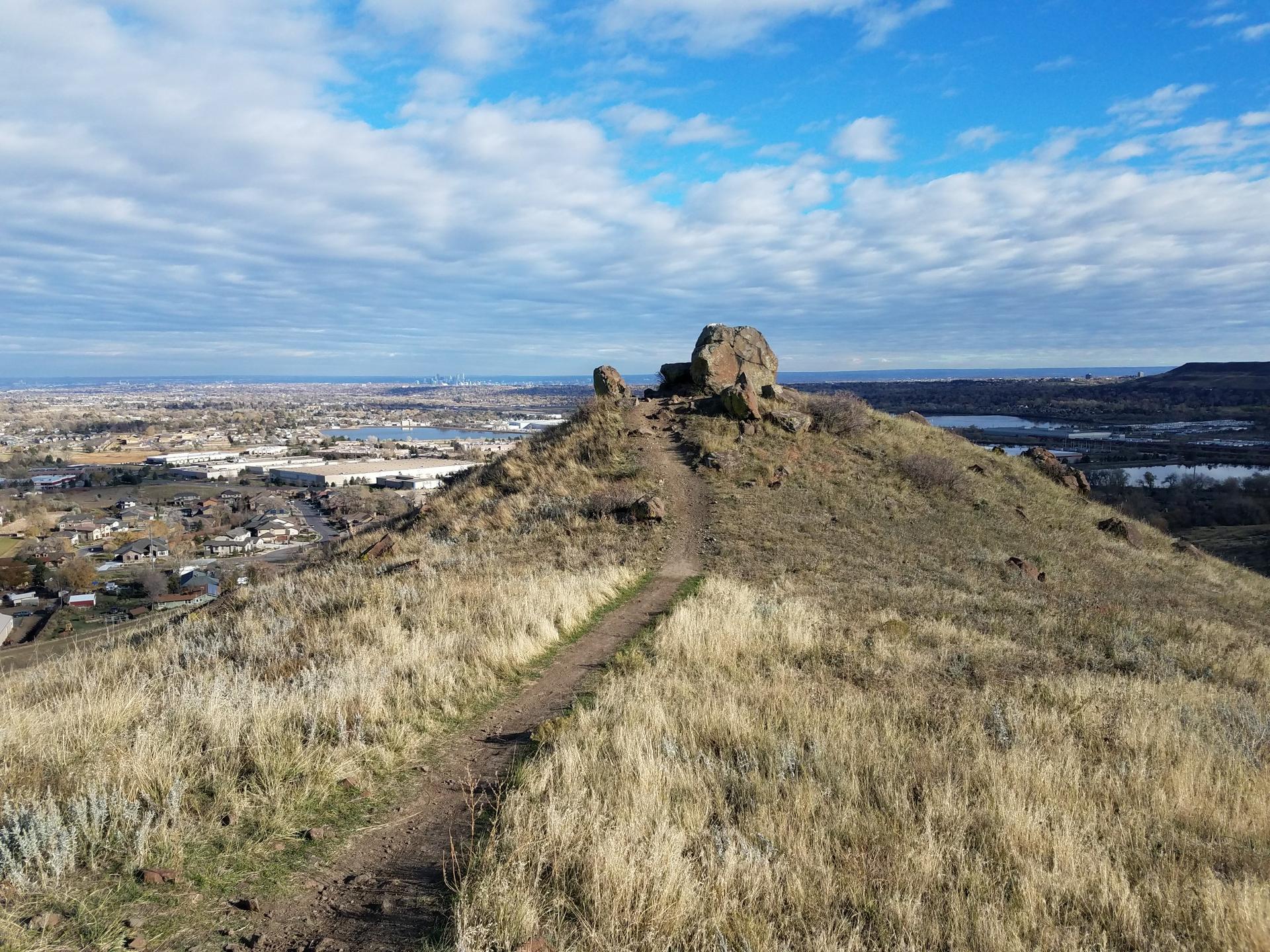 A dirt trail leads up a dry, rocky hill overlooking a distant city.