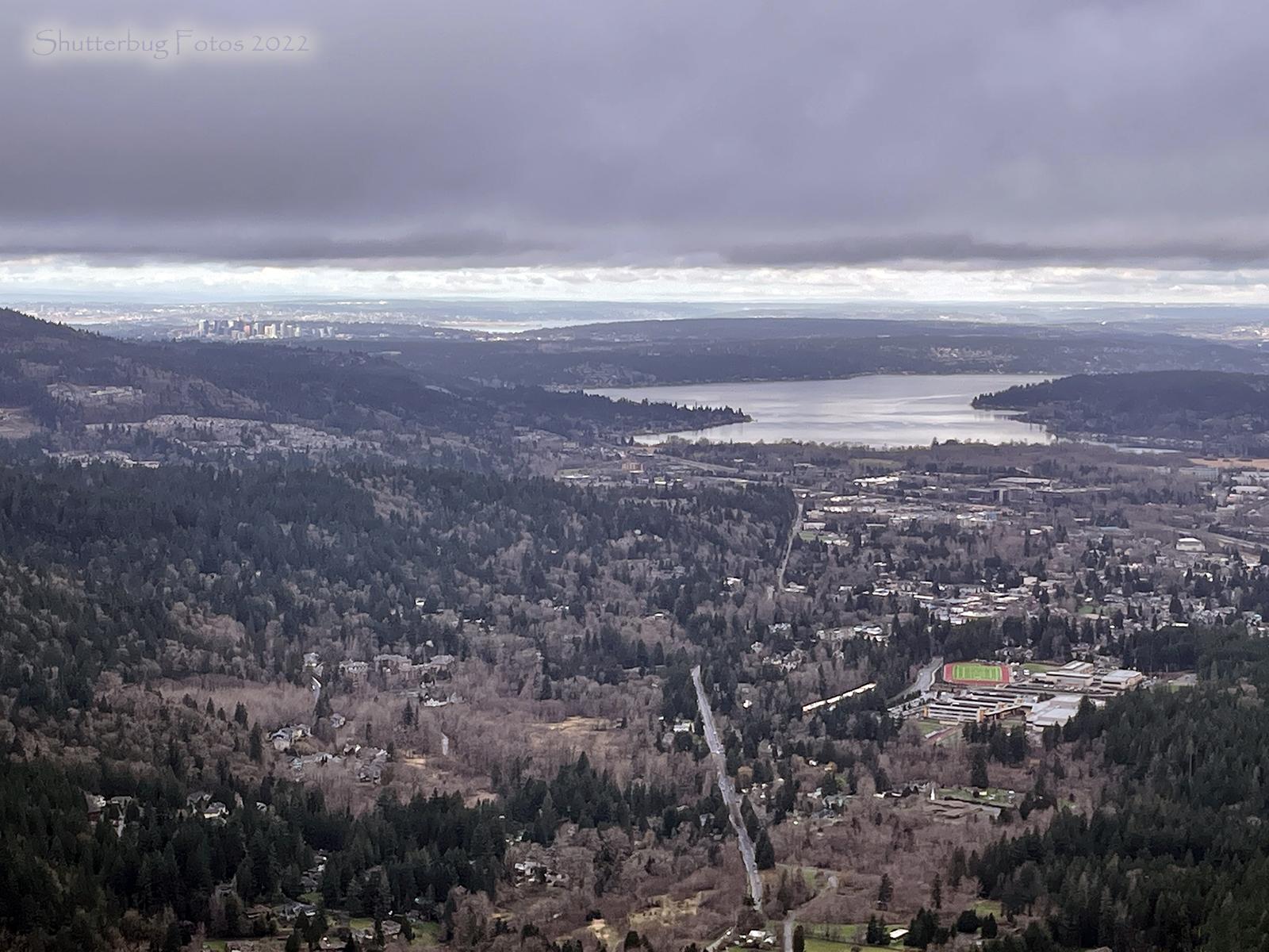 Aerial view of a lake, forested hills, and a distant city under cloudy skies.