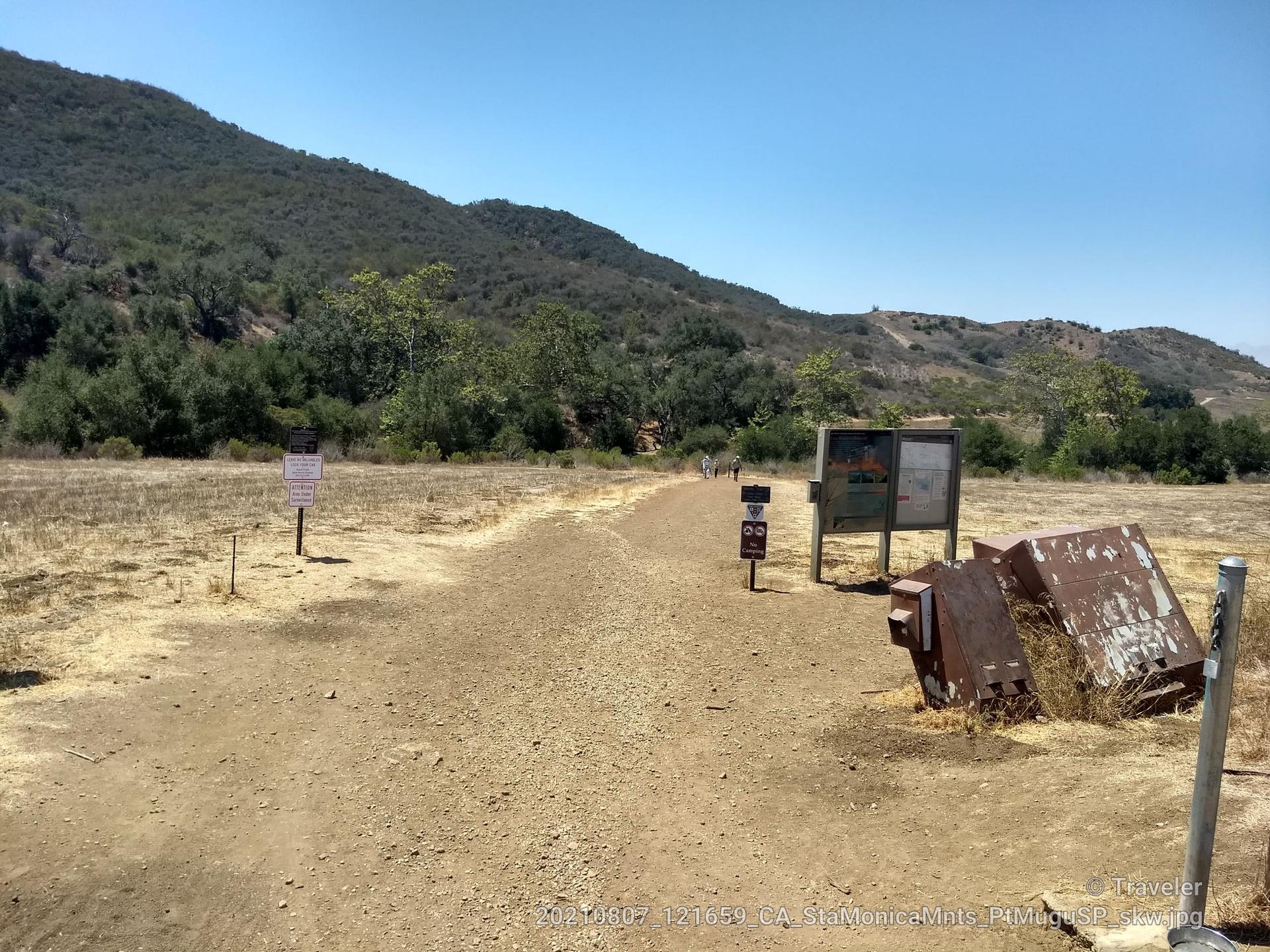 Wide dirt path with trail signs and hills under a bright, clear sky.