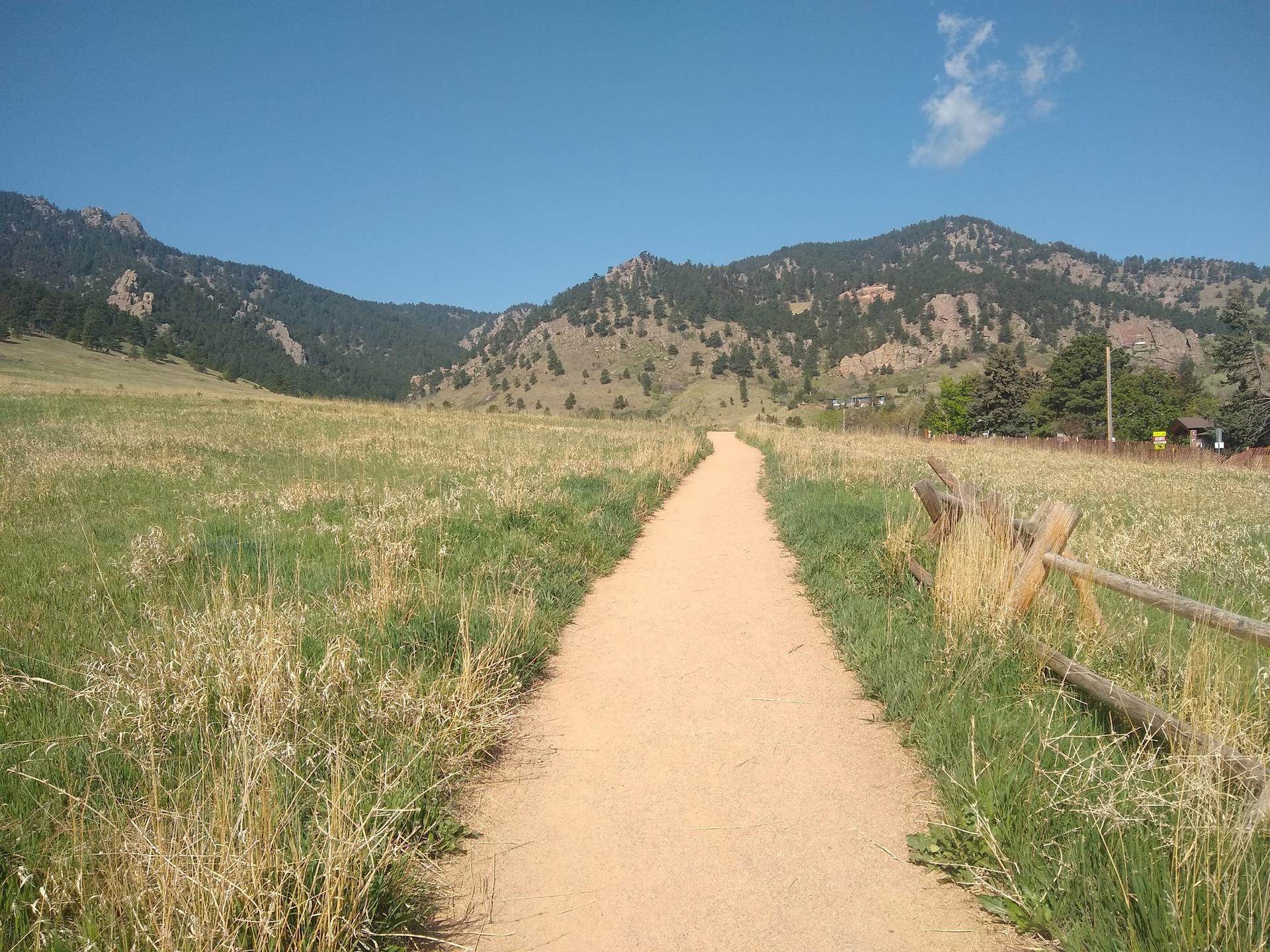 A dirt path leads through a sunny grassy field towards distant mountains.