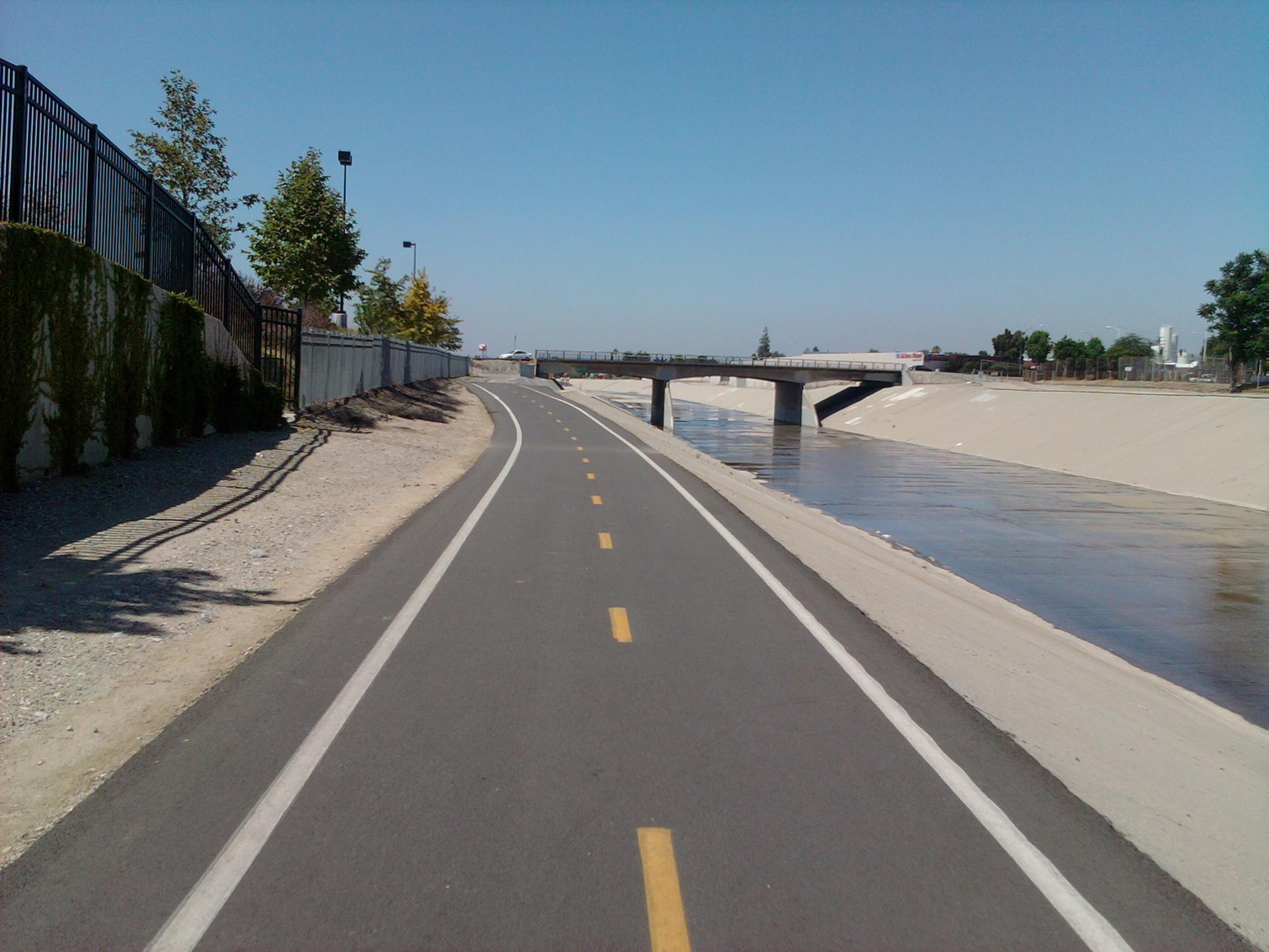 A paved path runs alongside a river with a bridge in the distance.
