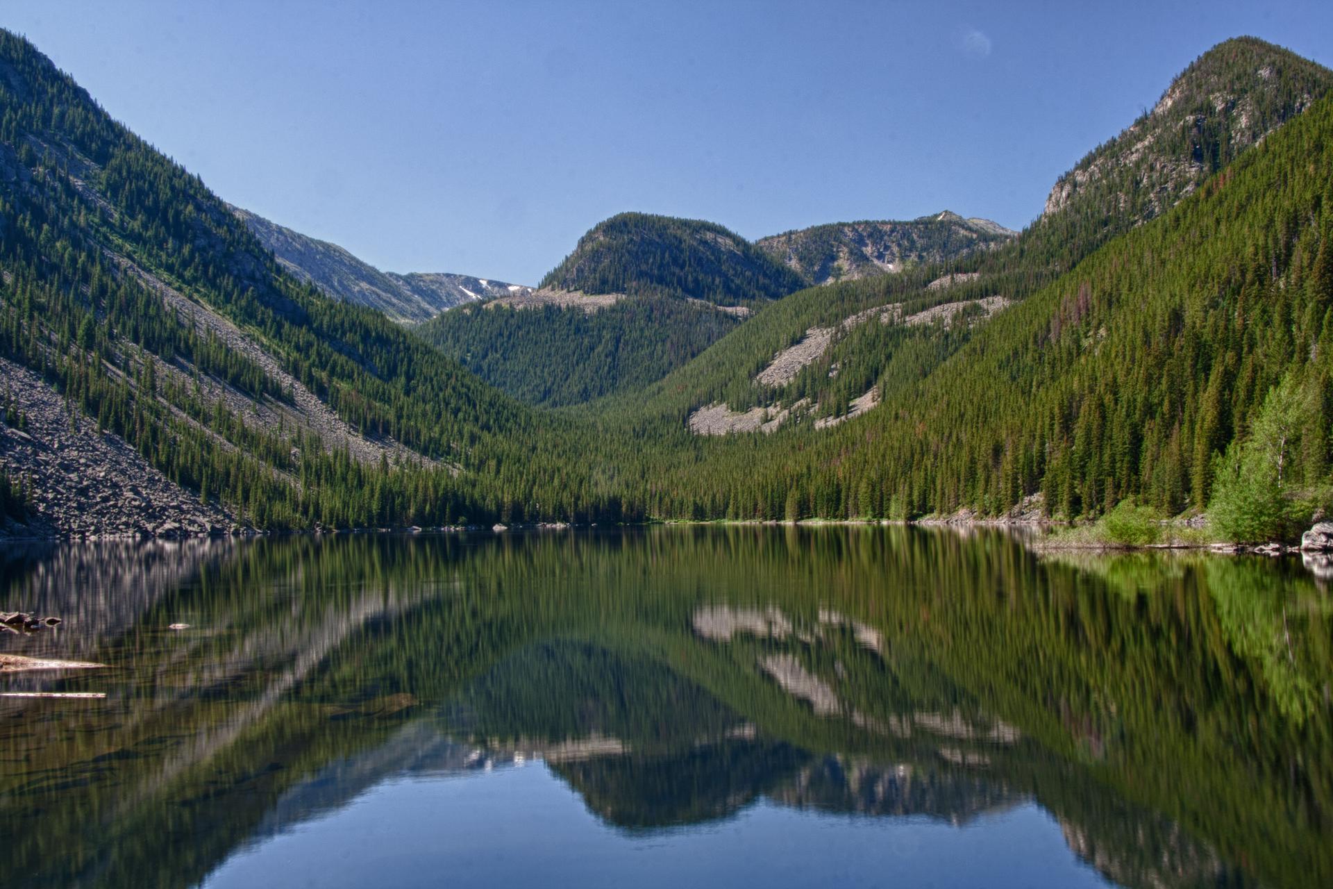Serene mountain lake reflecting steep, forested slopes under a clear blue sky.