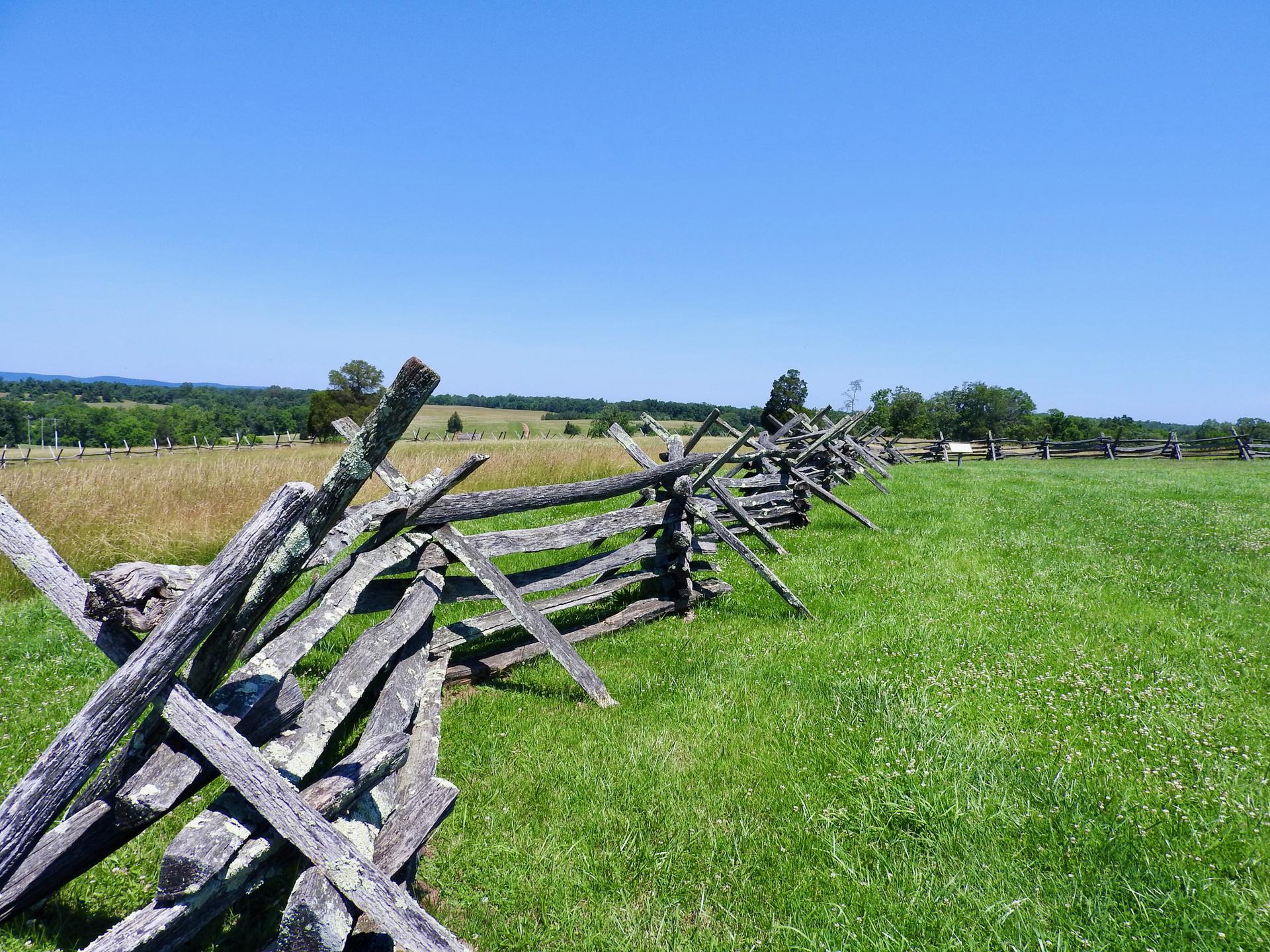 Weathered split-rail fence separates green and golden fields under a clear blue sky.