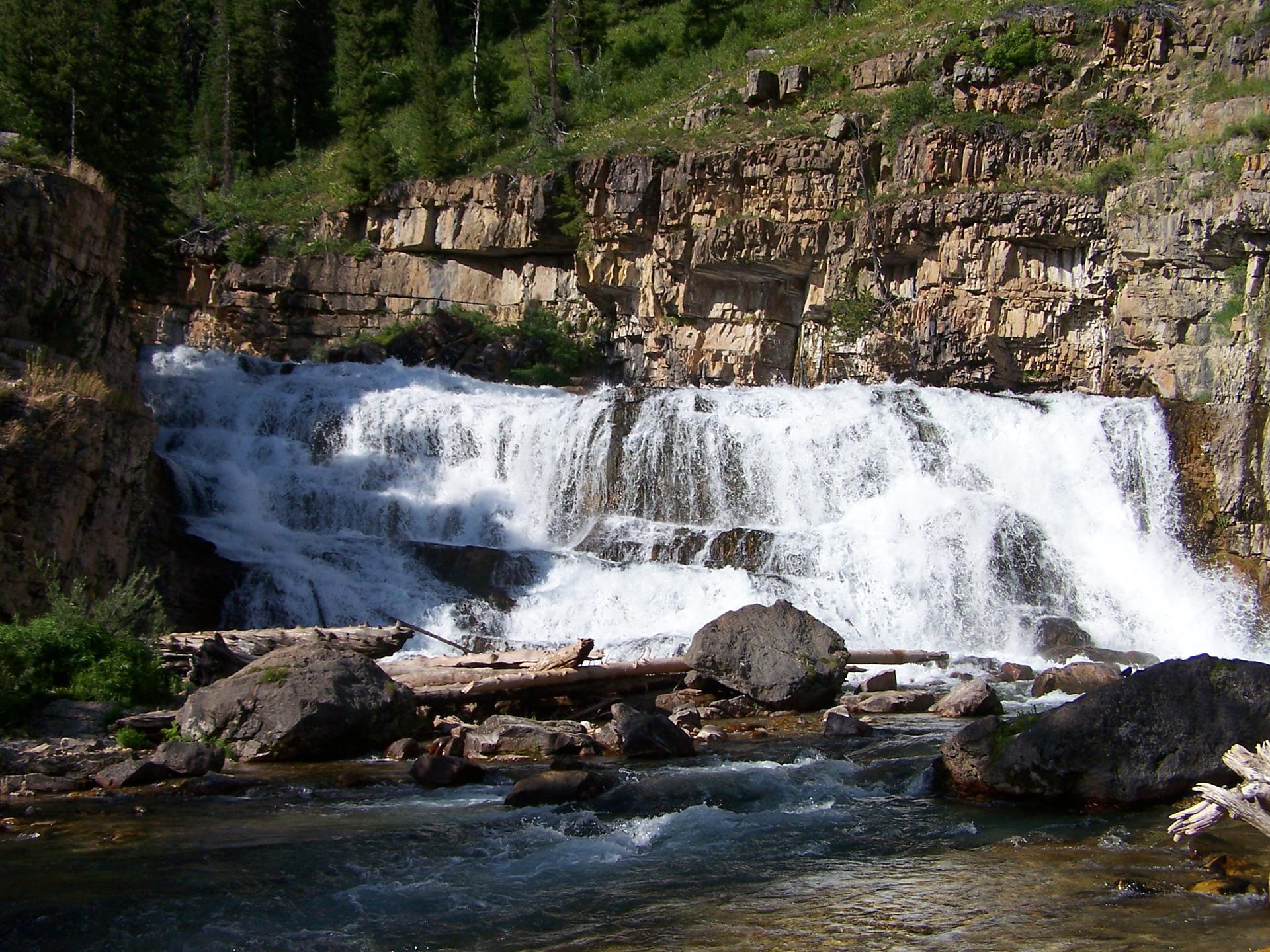 A powerful waterfall cascades over rocky ledges into a river under bright daylight.