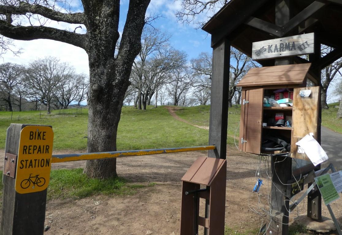 Bike repair station and wooden gate on a dirt path under a bright sky.