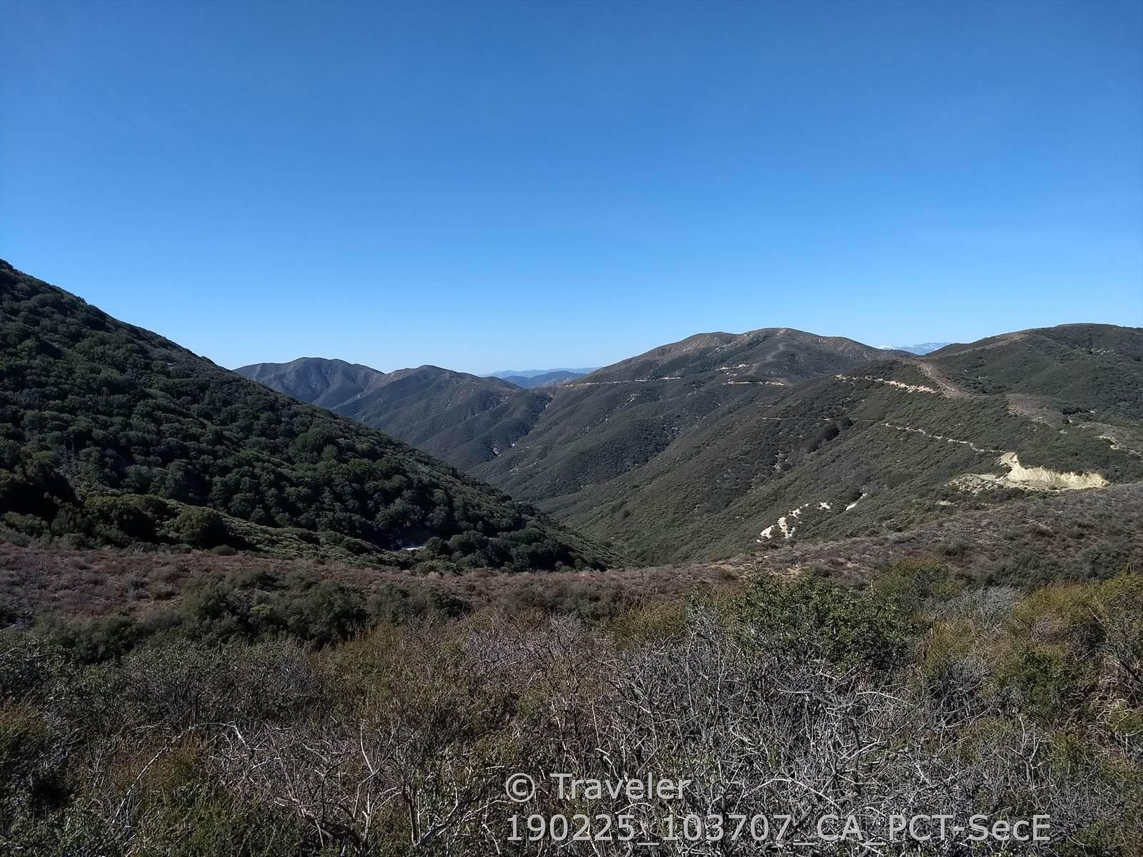 Vast mountainous landscape with green hills and a winding dirt road on a clear day.