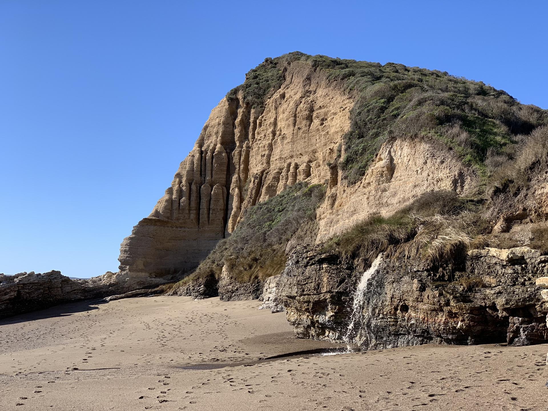 Sunny beach with a large sandy cliff and a small waterfall.