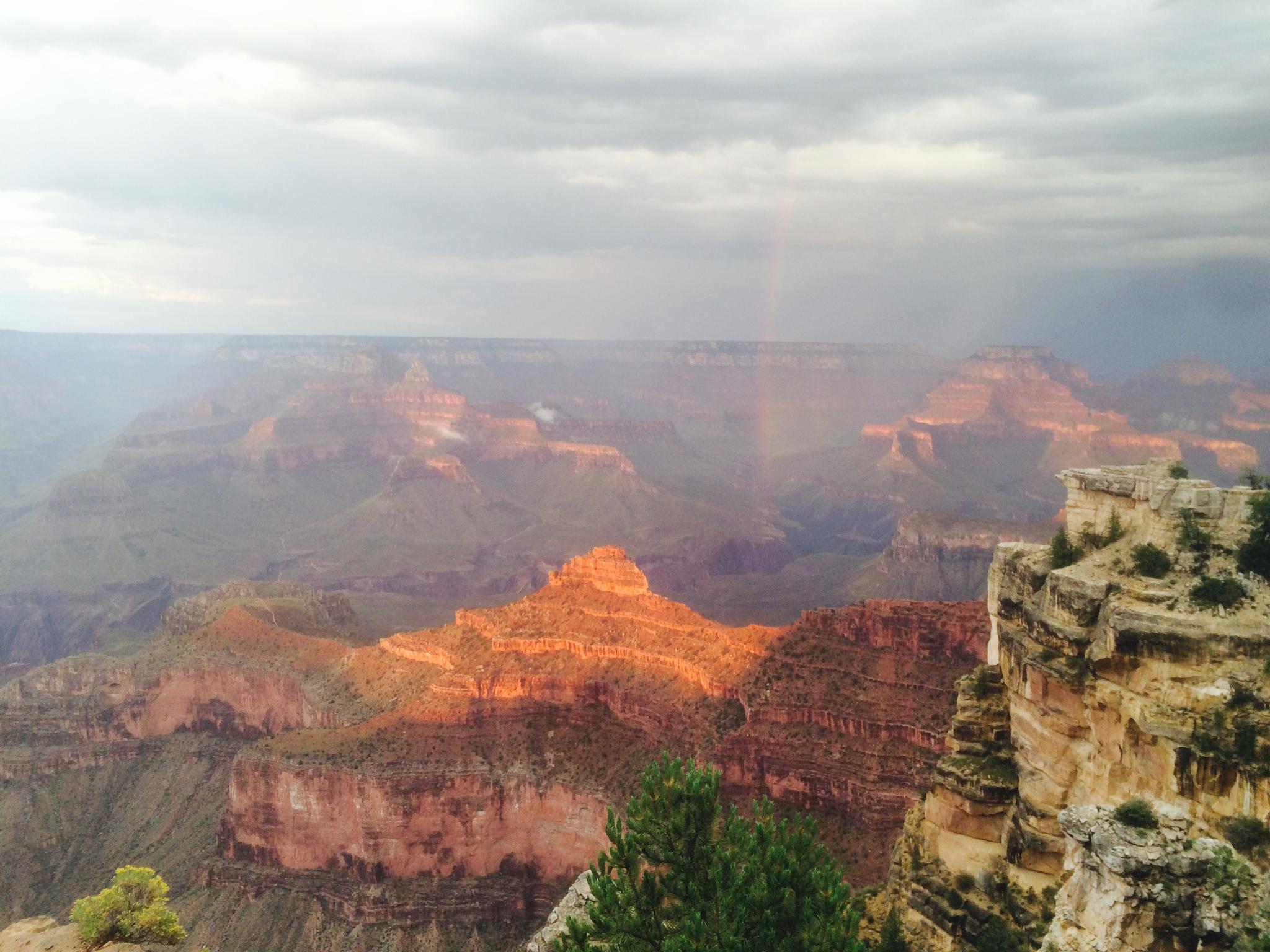 Expansive canyon landscape with a rainbow over sunlit red rock formations, cloudy sky.