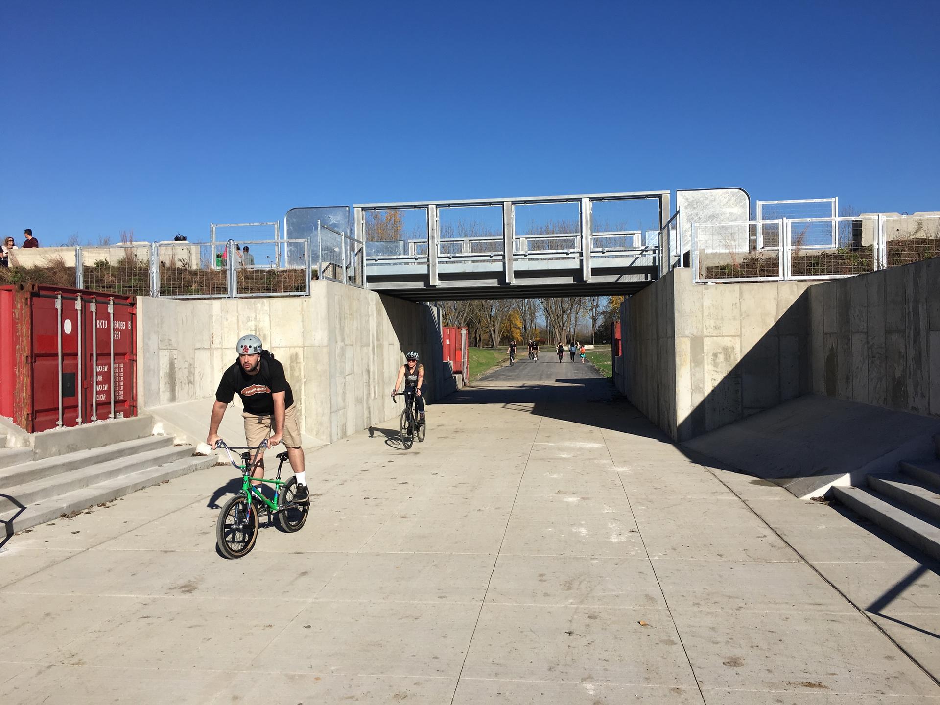 People cycling on a concrete path under a bridge on a bright, sunny day.