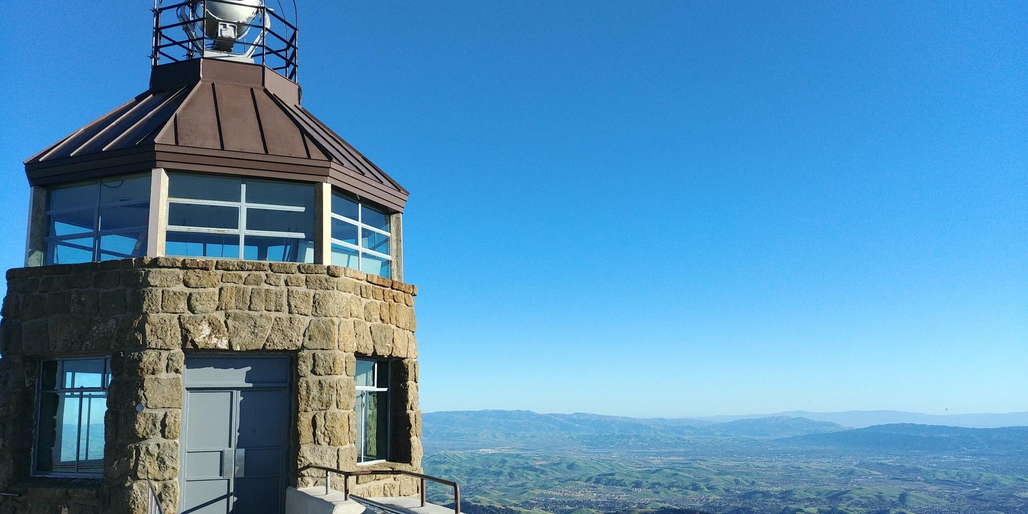 Stone tower with a metal roof against a clear blue sky and distant hills.