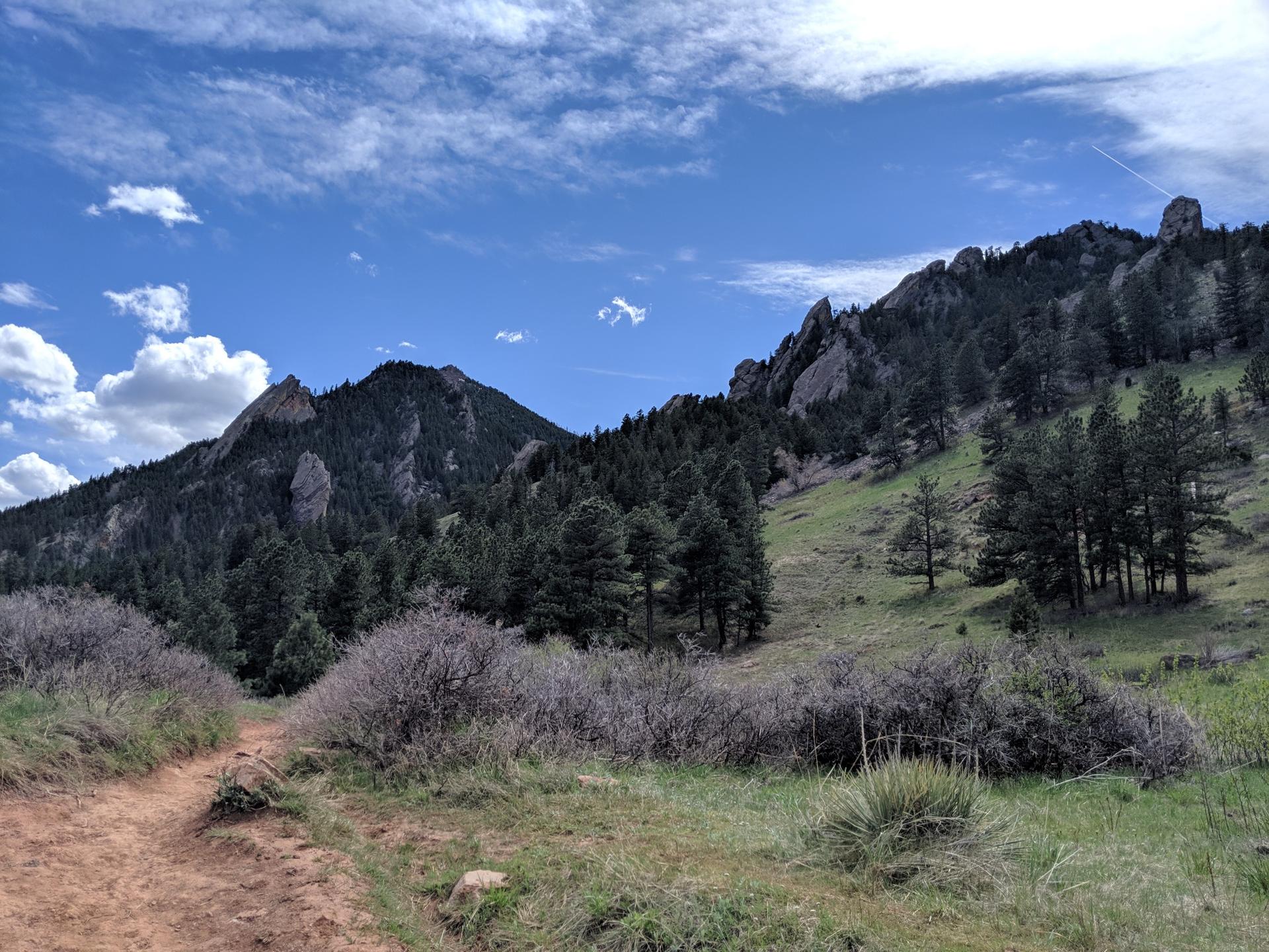 Dirt path leading towards green hills and rugged mountains under a cloudy sky.