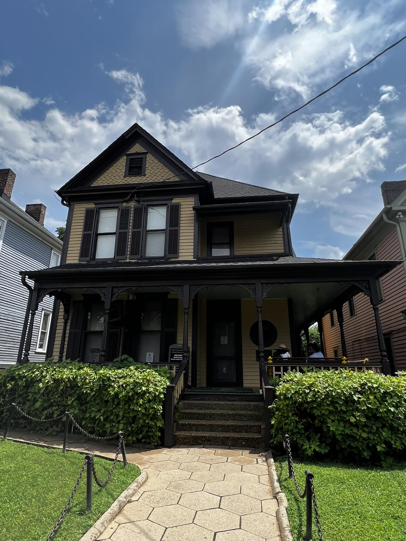 Historic yellow house with dark trim and a porch under a partly cloudy sky.