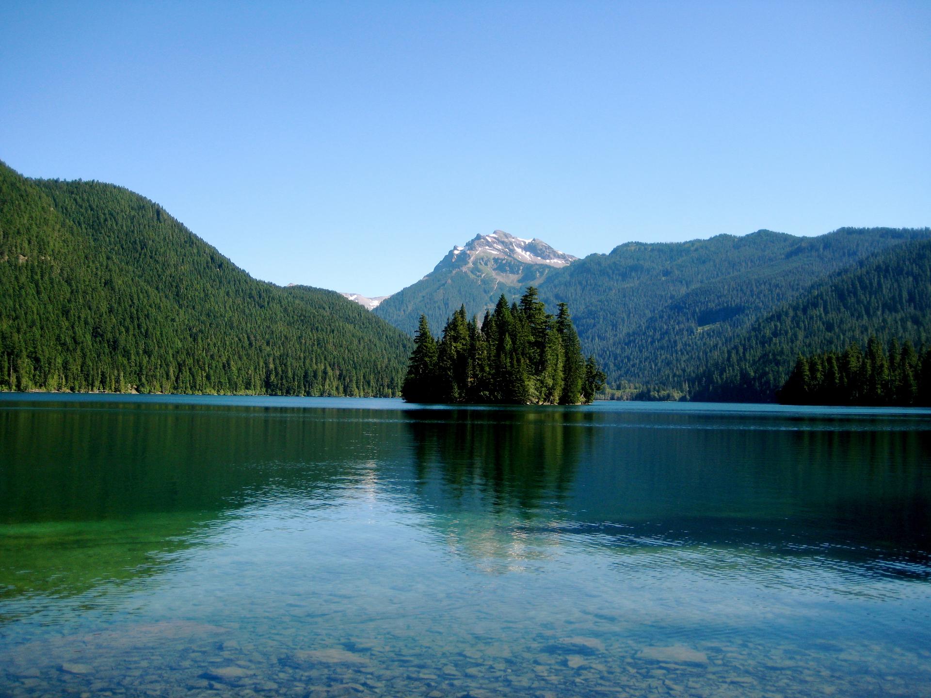 Serene mountain lake with a tree-covered island, reflecting blue sky and distant peaks.