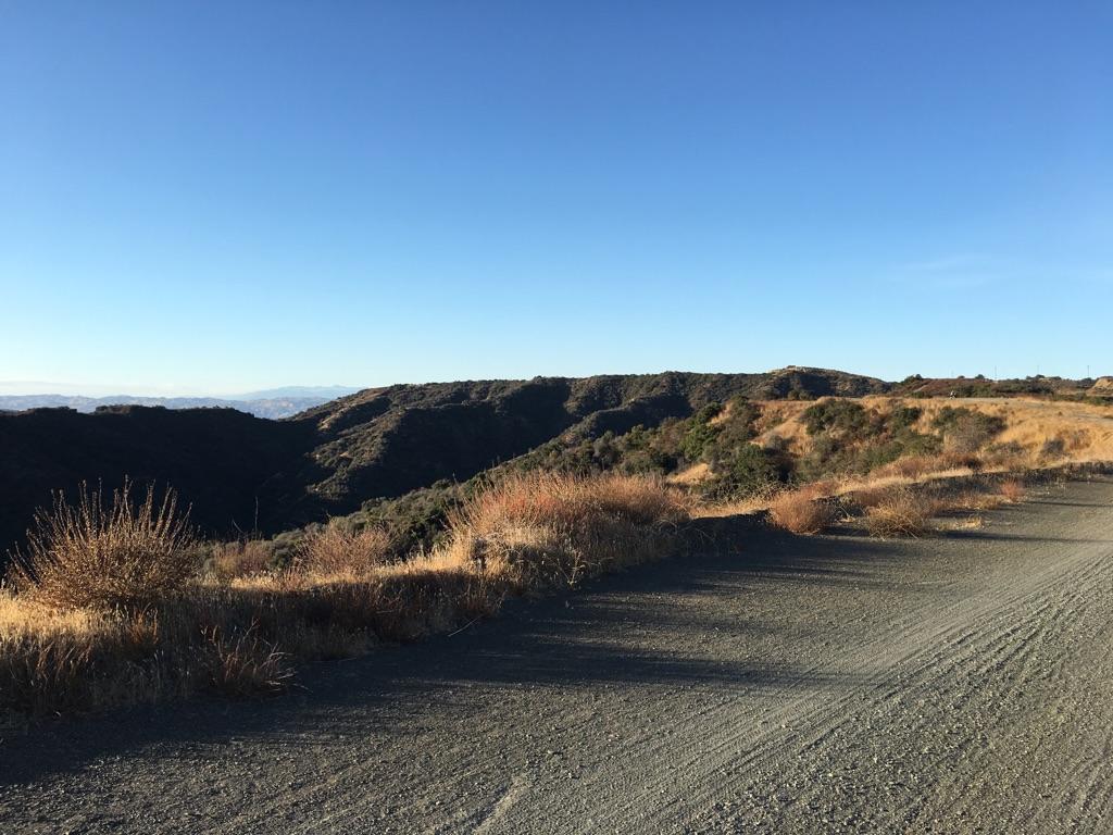 Gravel road on a hillside overlooking green and brown mountains under clear blue sky.