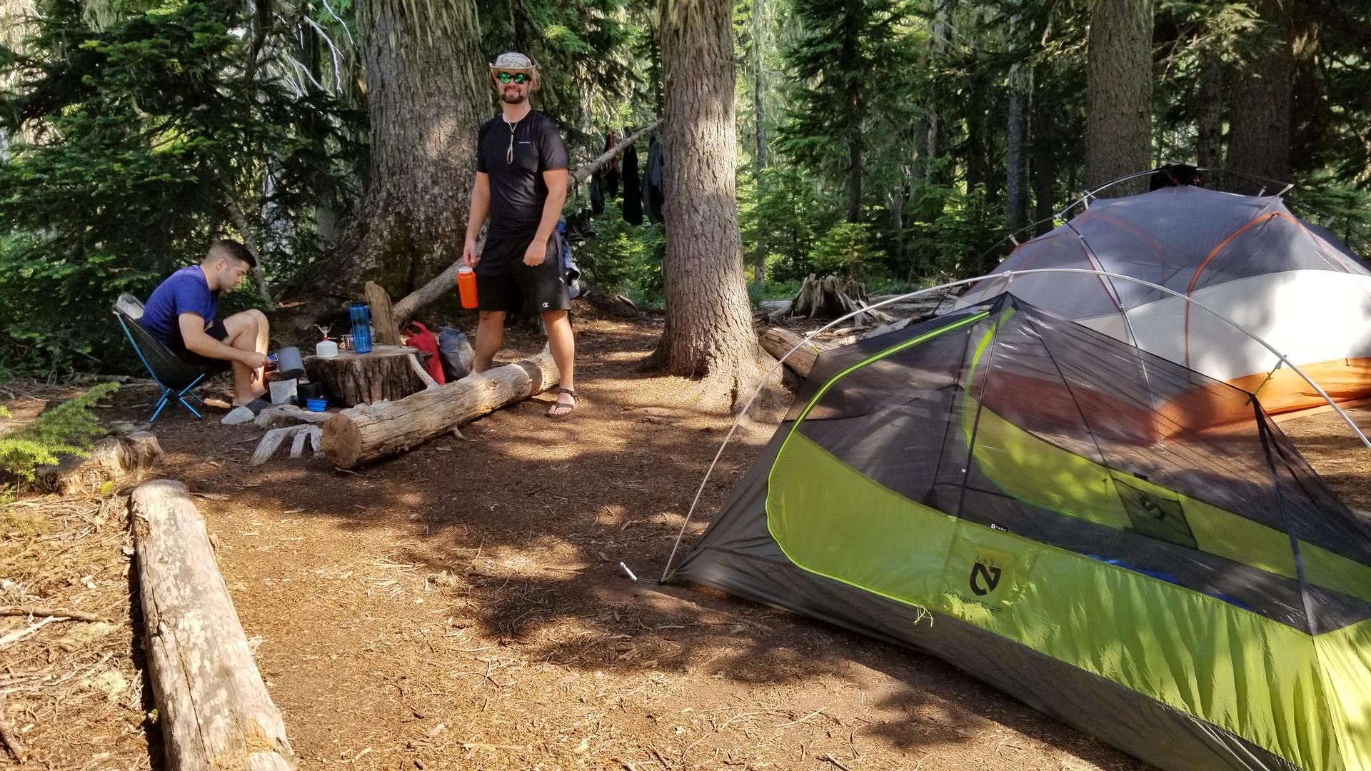 Two men camping in a forest with a tent and cooking gear.