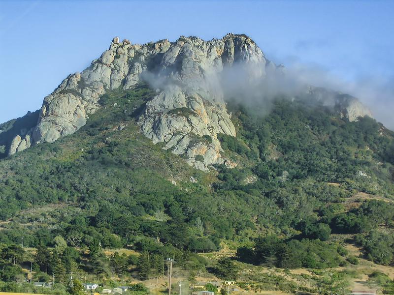 Majestic rocky mountain peak with green trees, partly shrouded in mist, under clear sky.