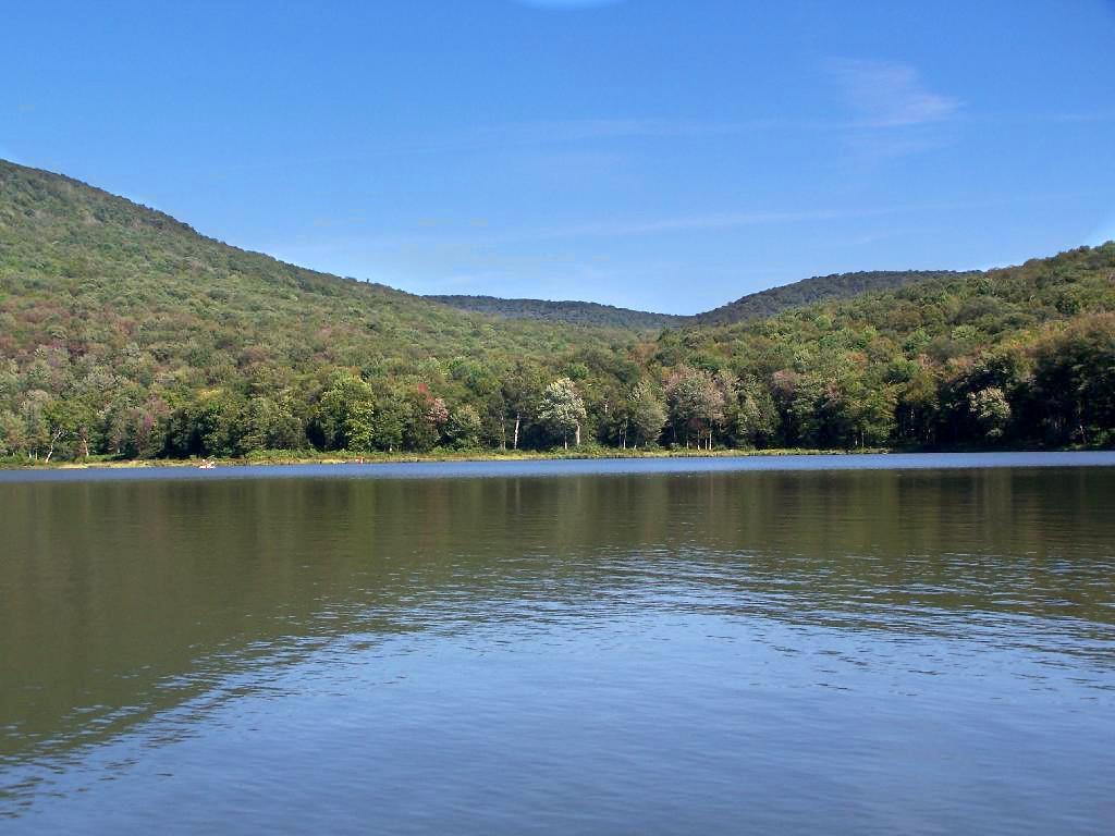 Calm lake reflecting forested hills under a bright blue sky.