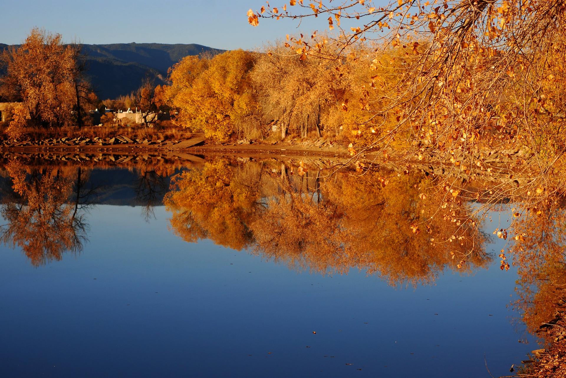 Calm lake reflecting golden autumn trees and mountains under clear sky.