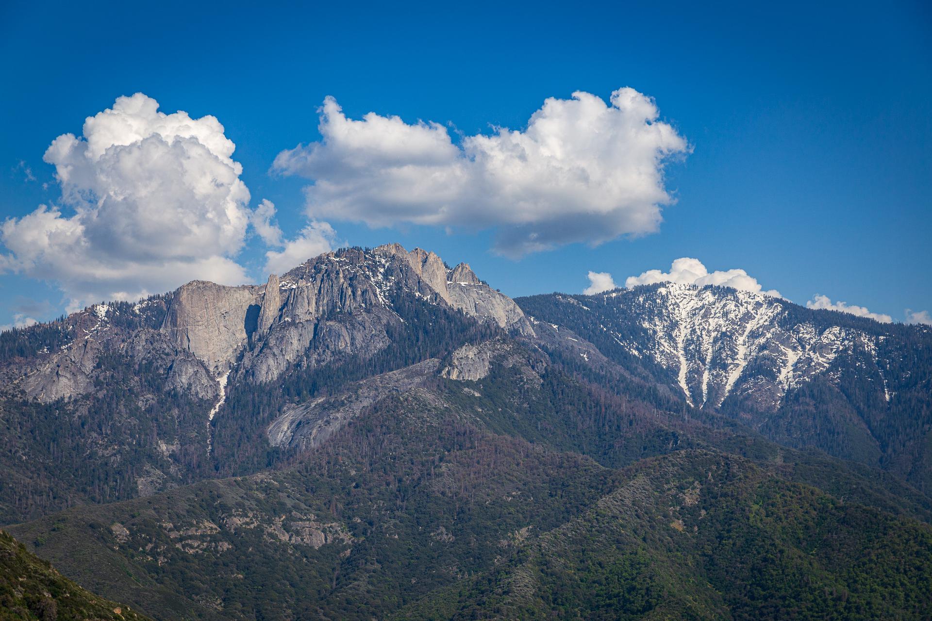 Rugged mountain peaks, some snow-capped, under a bright blue sky with clouds.