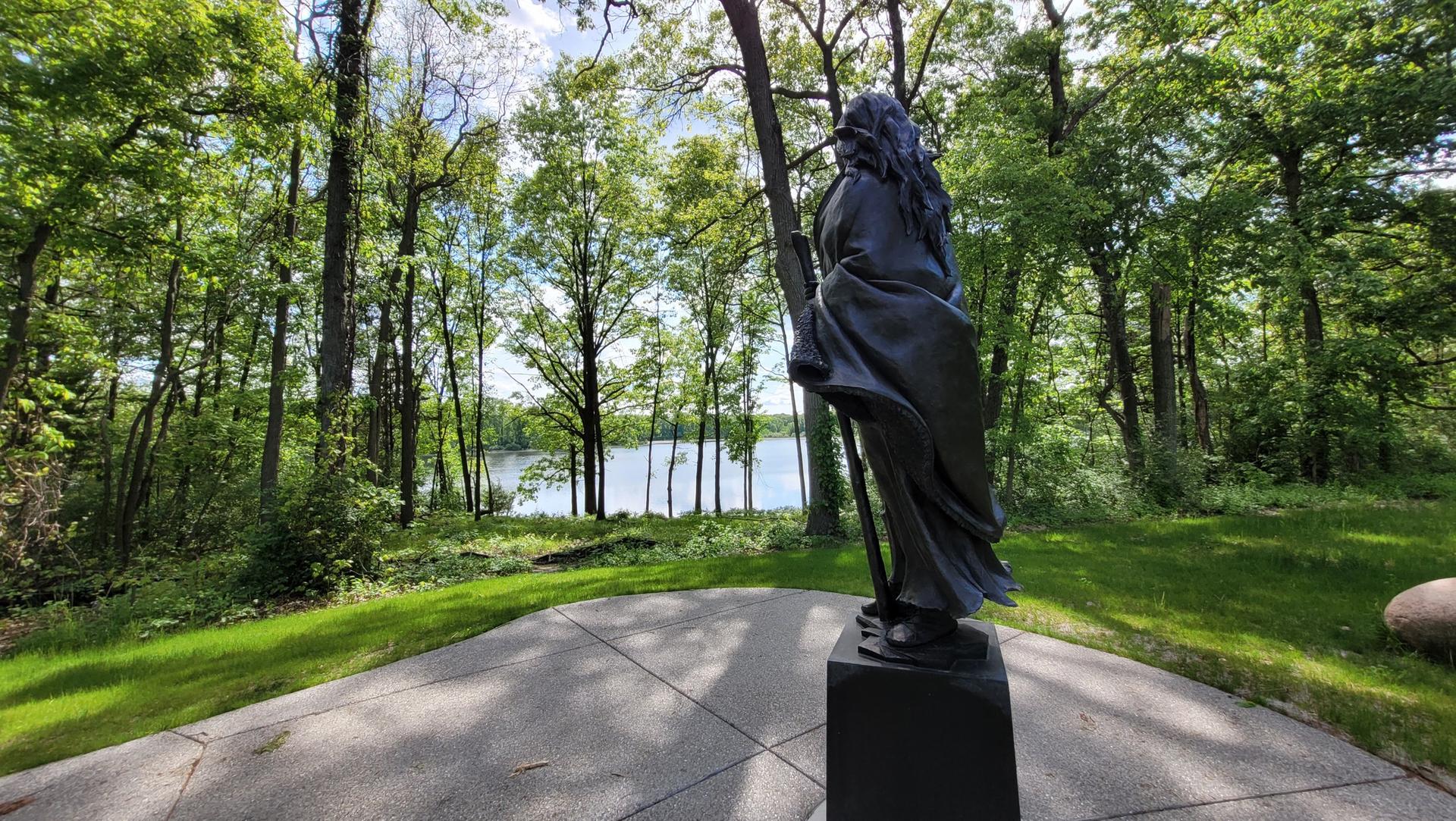 A dark statue of a robed figure overlooks a lake through green trees.