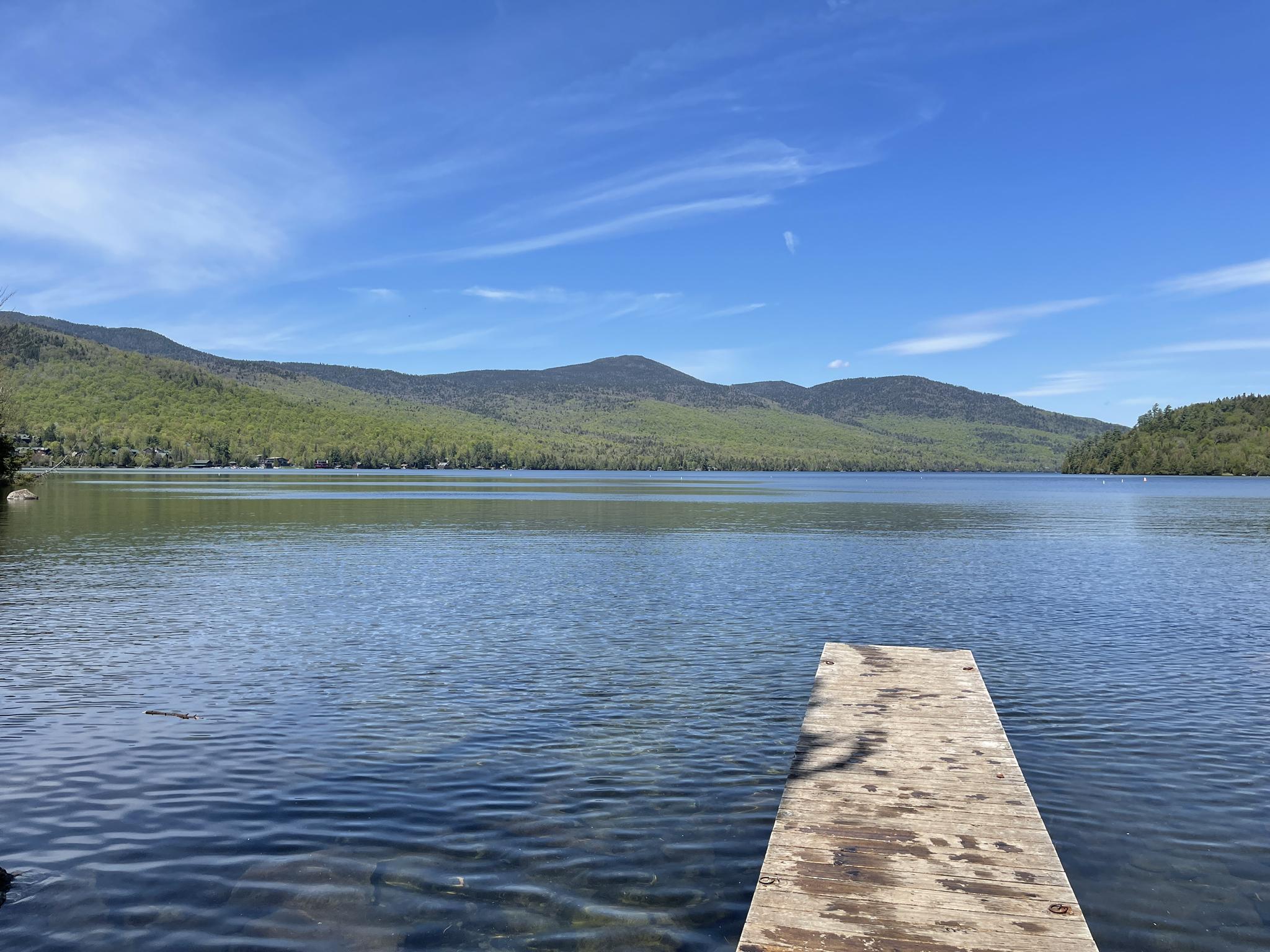 Wooden dock extends into a calm lake with forested mountains under a blue sky.