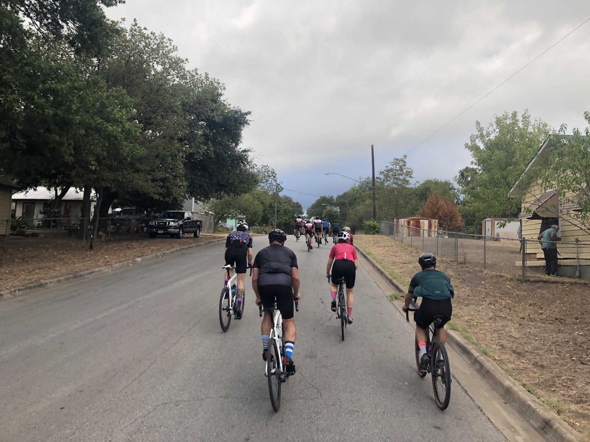 A group of cyclists rides down a paved road under a cloudy sky.