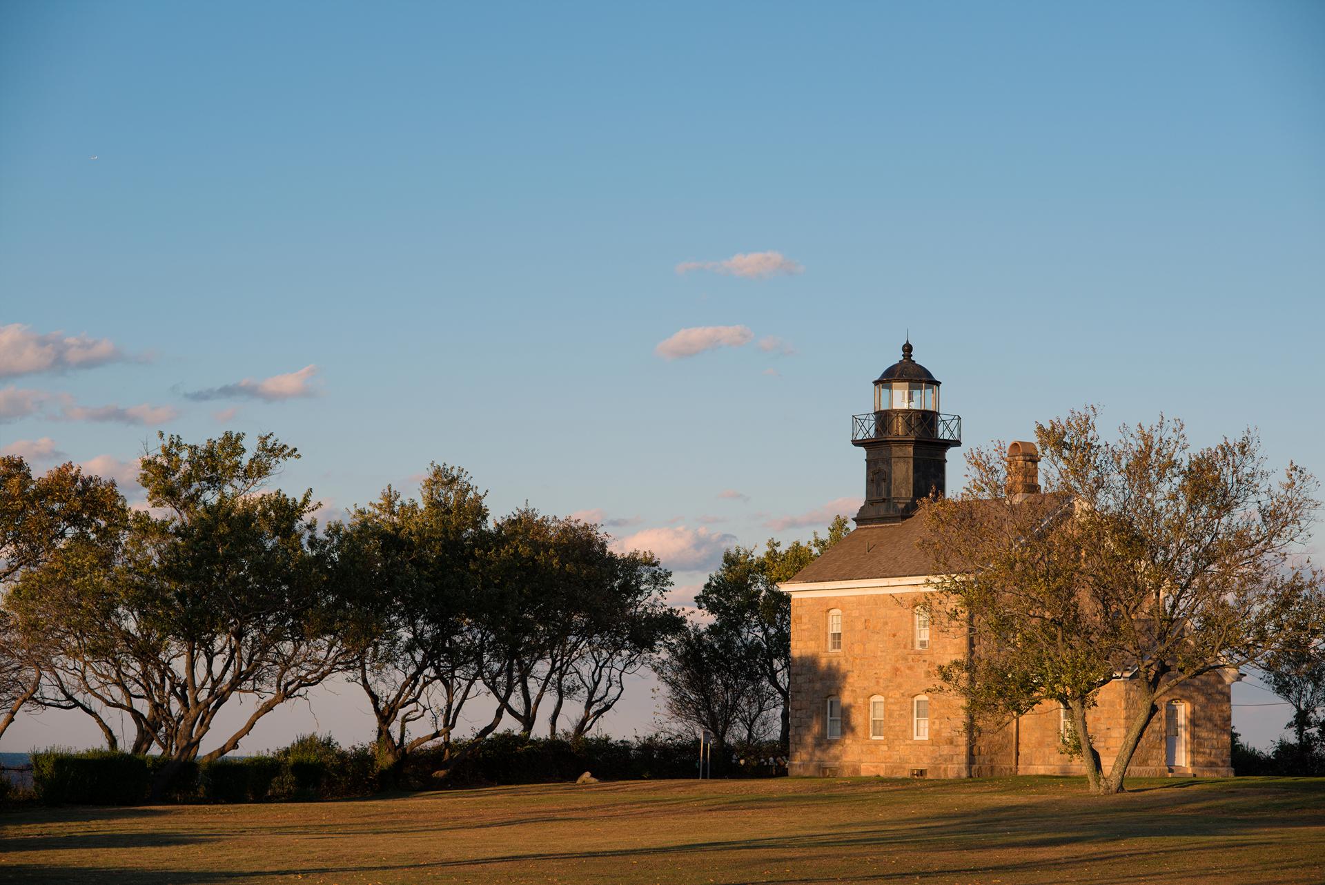 Historic lighthouse and building in warm evening light beside trees.