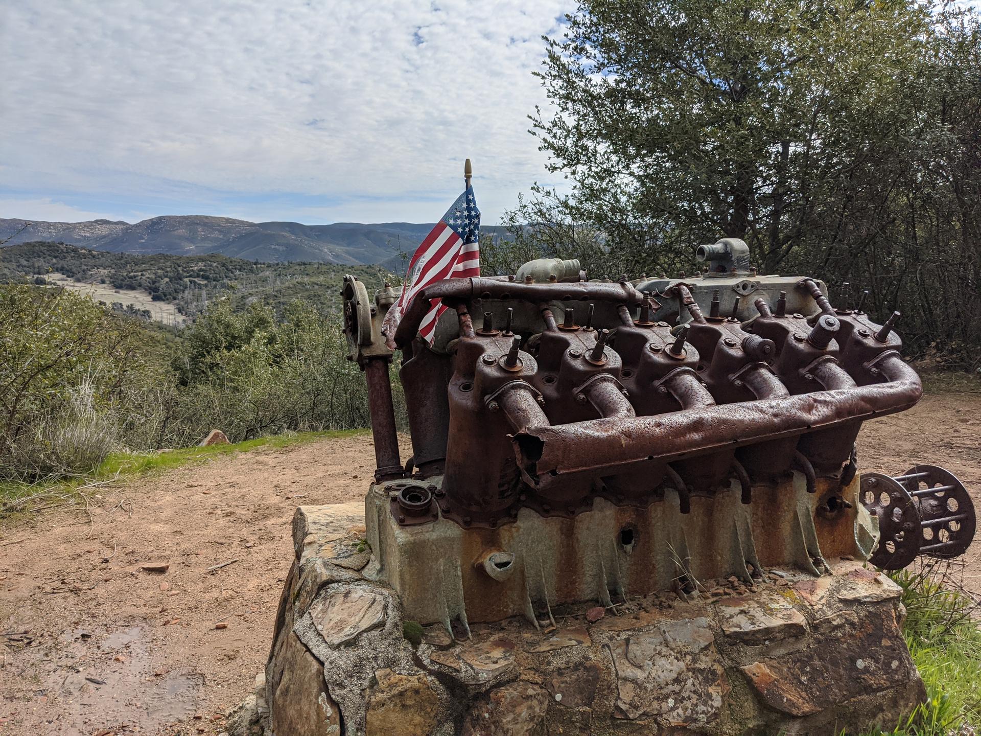 Rusted engine block with American flag overlooks a hilly, green landscape.