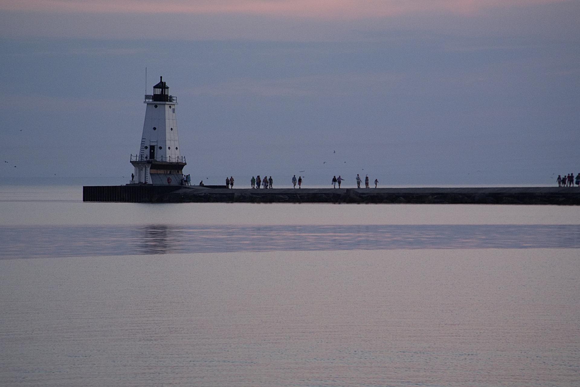 A white lighthouse on a pier with people walking at dusk.