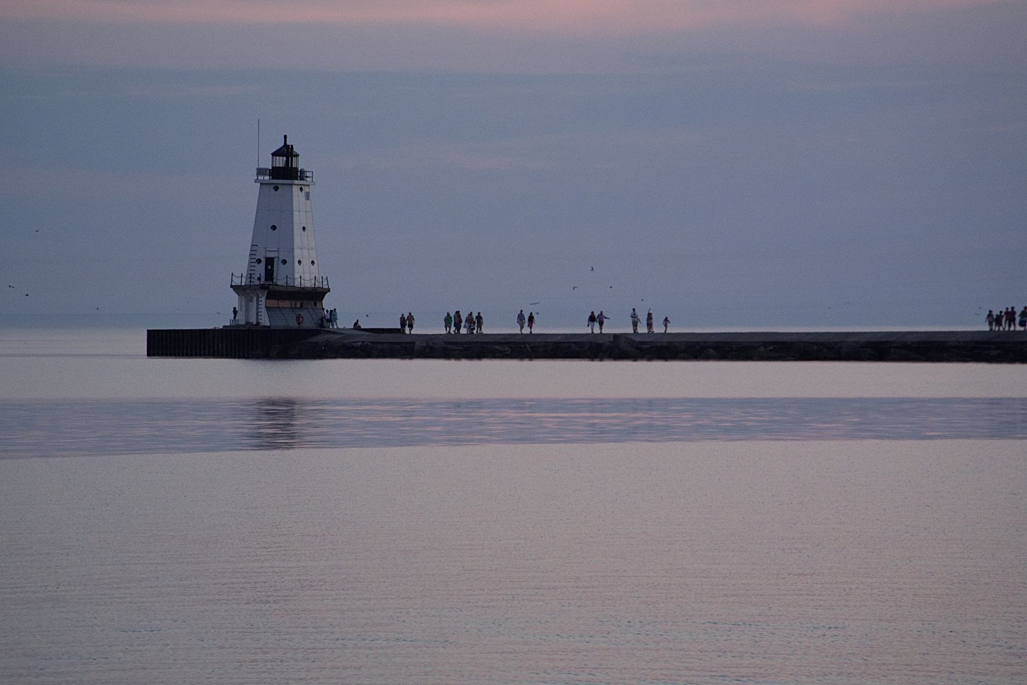 A white lighthouse on a pier with people walking at dusk. - Ludington North Breakwater Trail