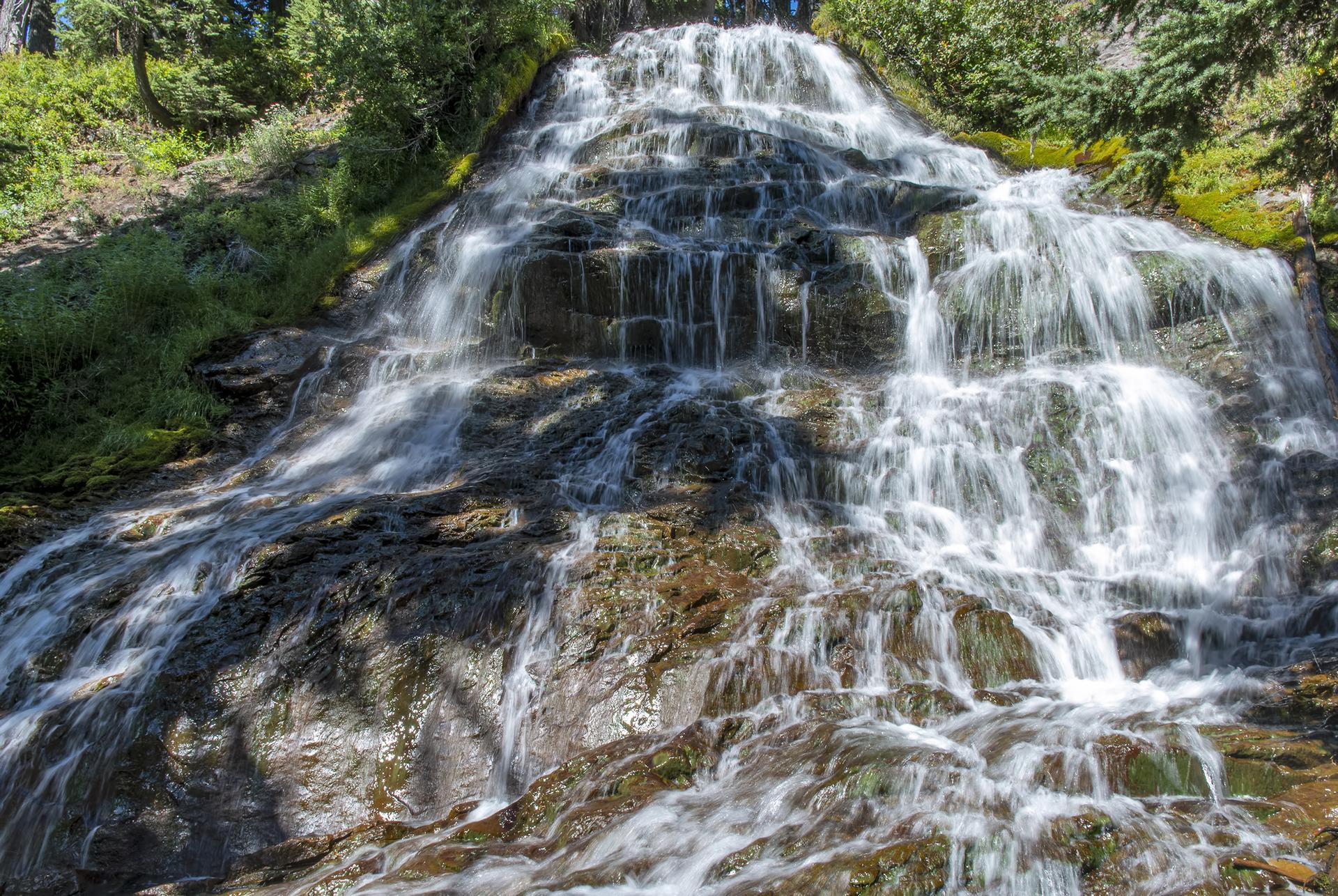 A multi-tiered waterfall cascades over dark rocks, surrounded by green foliage.