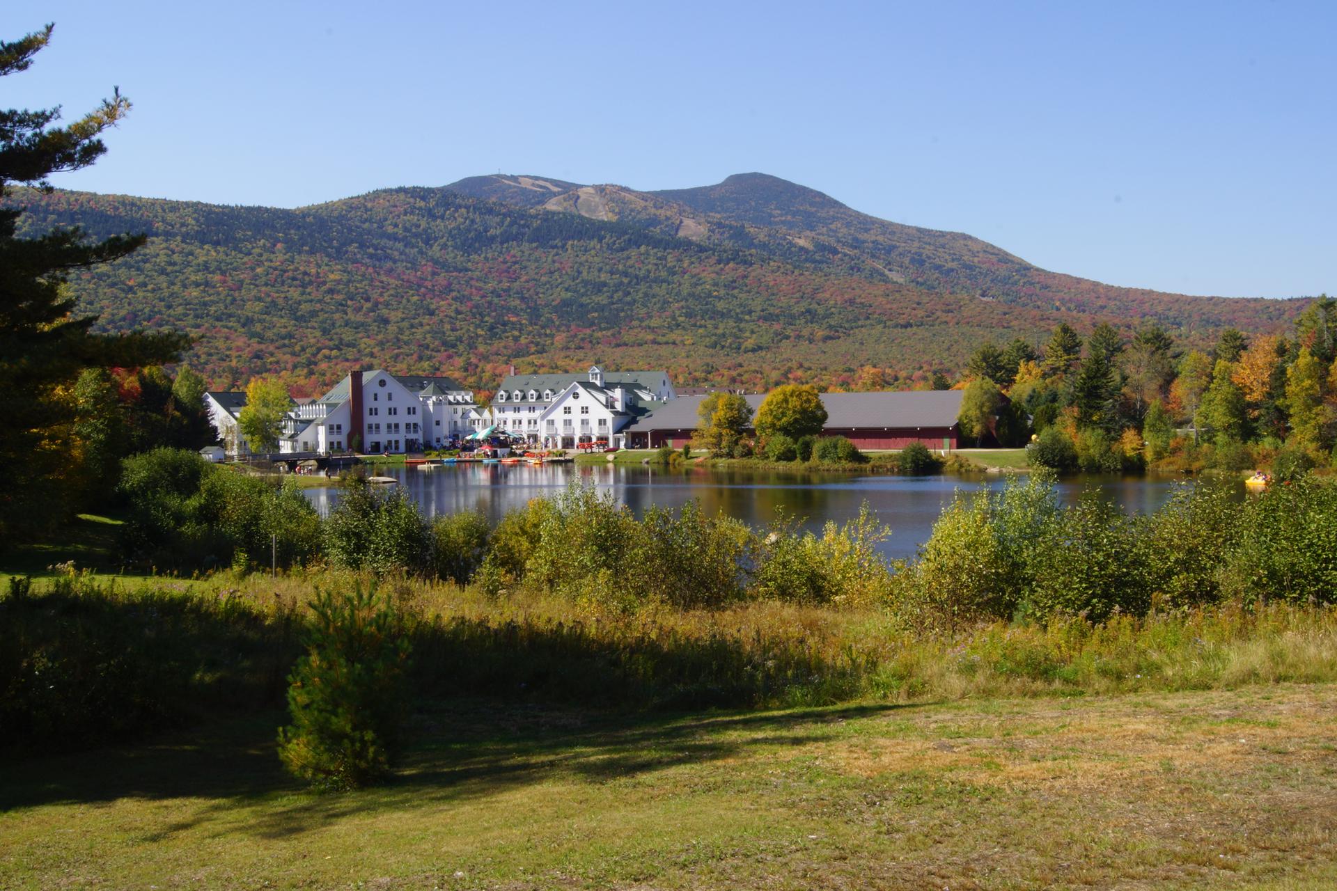 White buildings by a lake with autumn trees and a mountain under sunny skies.