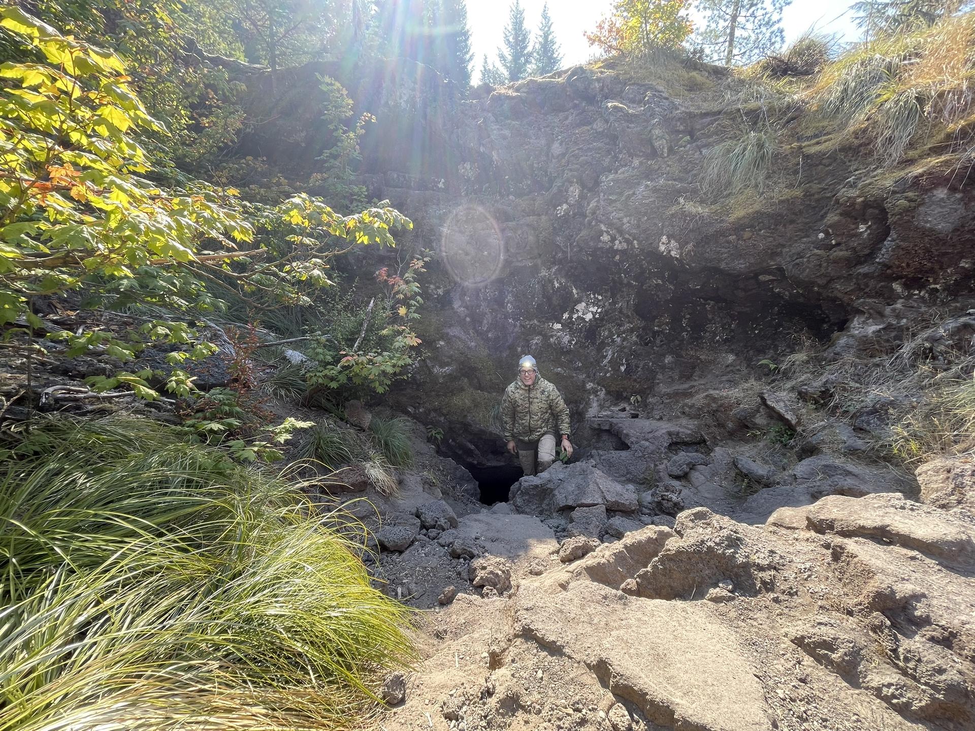 Person with headlamp entering a dark cave opening on a sunny day.