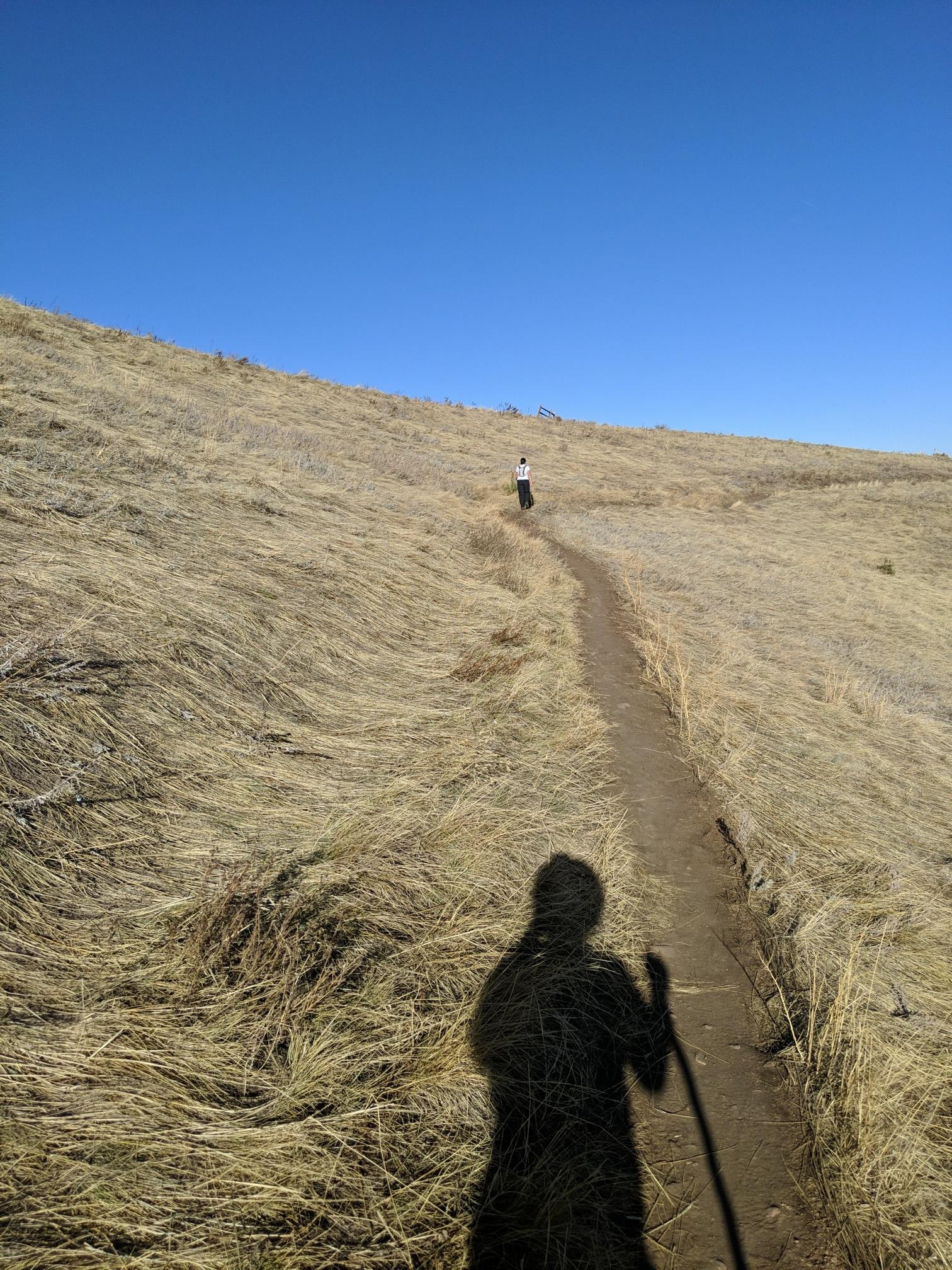Hiker's shadow on a dry, grassy trail leading to another distant hiker.