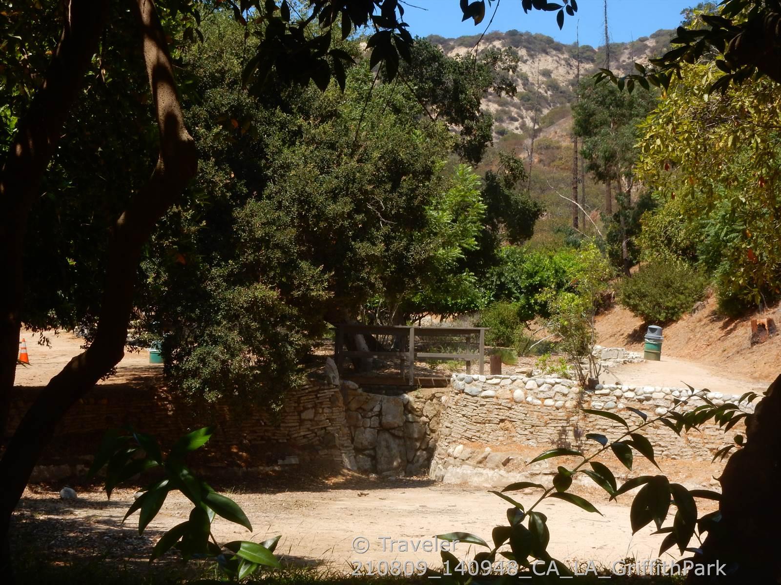Sunny wooded path with stone walls, green trees, and distant hills.