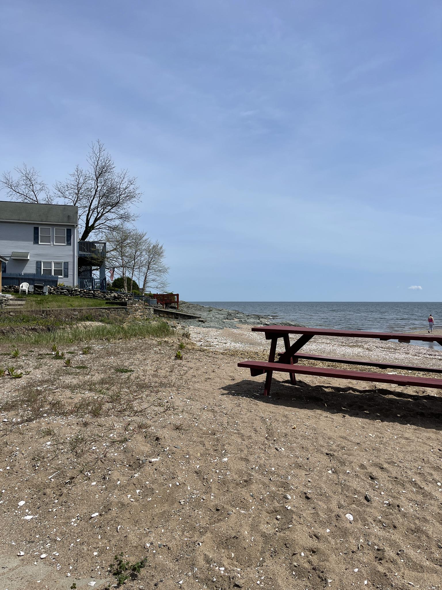 A red picnic table sits on a sandy beach next to the ocean and a house.