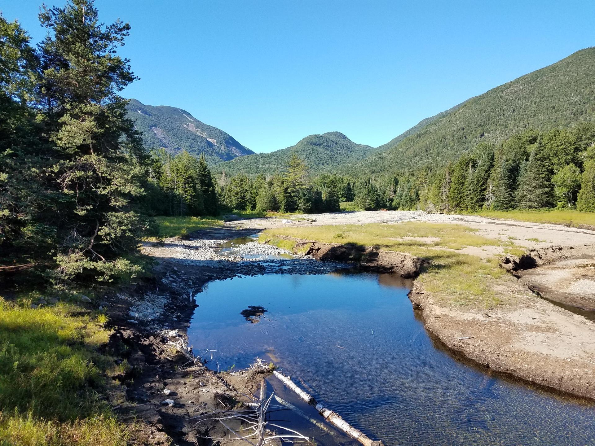 A clear river flowing through a valley with forested mountains under a blue sky.