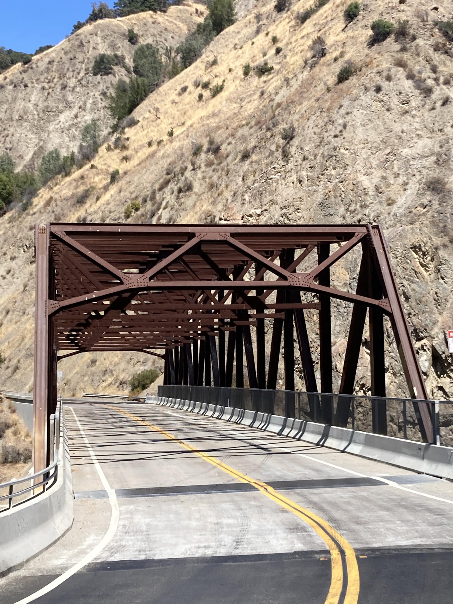 A brown metal truss bridge spans a road next to a dry, rocky hillside.